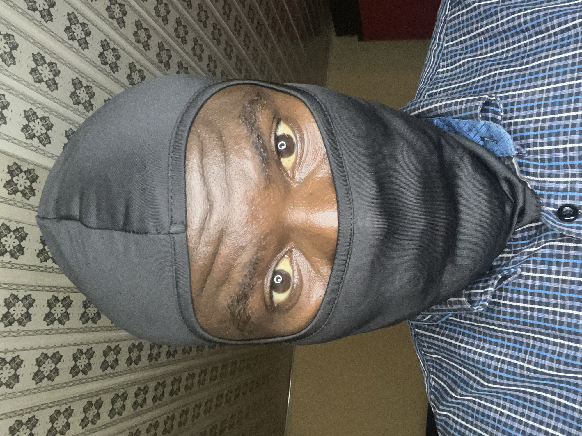 A person wearing a black balaclava is facing the camera. The person has dark skin and brown eyes. They are wearing a blue and white checkered shirt. The background includes a wall with patterned wallpaper. The location is Gura Top, Nigeria.