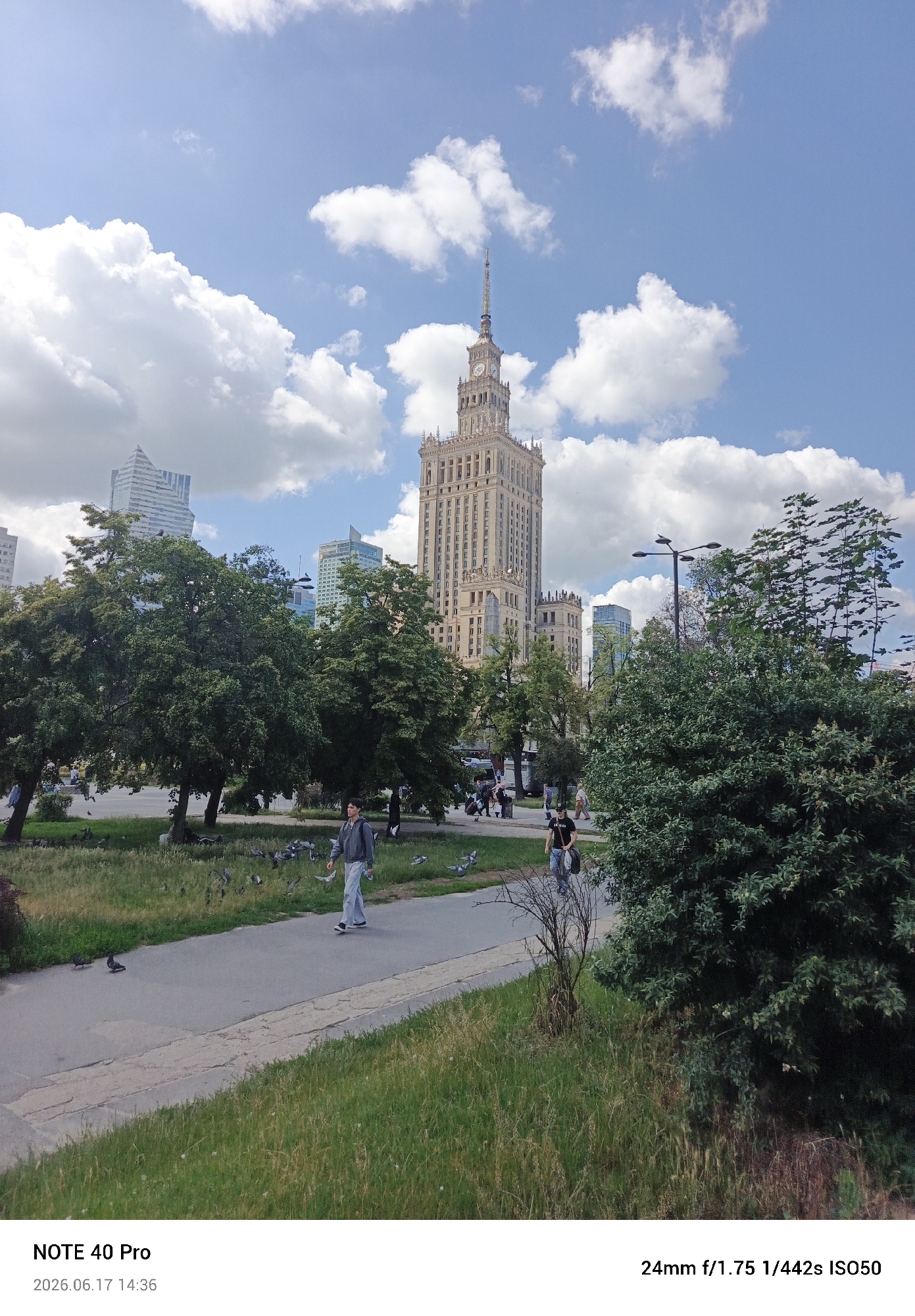 The image depicts the Palace of Culture and Science in Warsaw, Poland, on a sunny day with scattered clouds. A paved pathway curves through a grassy park in the foreground, where several pigeons are visible. Two individuals are walking on the pathway: one in the middle ground, wearing a gray hooded jacket and light-colored pants, is walking towards the left, and another person, wearing a dark t-shirt and carrying bags, is walking in the background towards the right. In the mid-ground, large, leafy trees are prominent, with the iconic Palace of Culture and Science towering behind them. Several other modern buildings are visible in the background, forming a city skyline. A lamppost with a bird on top is also present in the mid-ground. The timestamp at the bottom indicates the photo was taken on June 17, 2026, at 14:36.