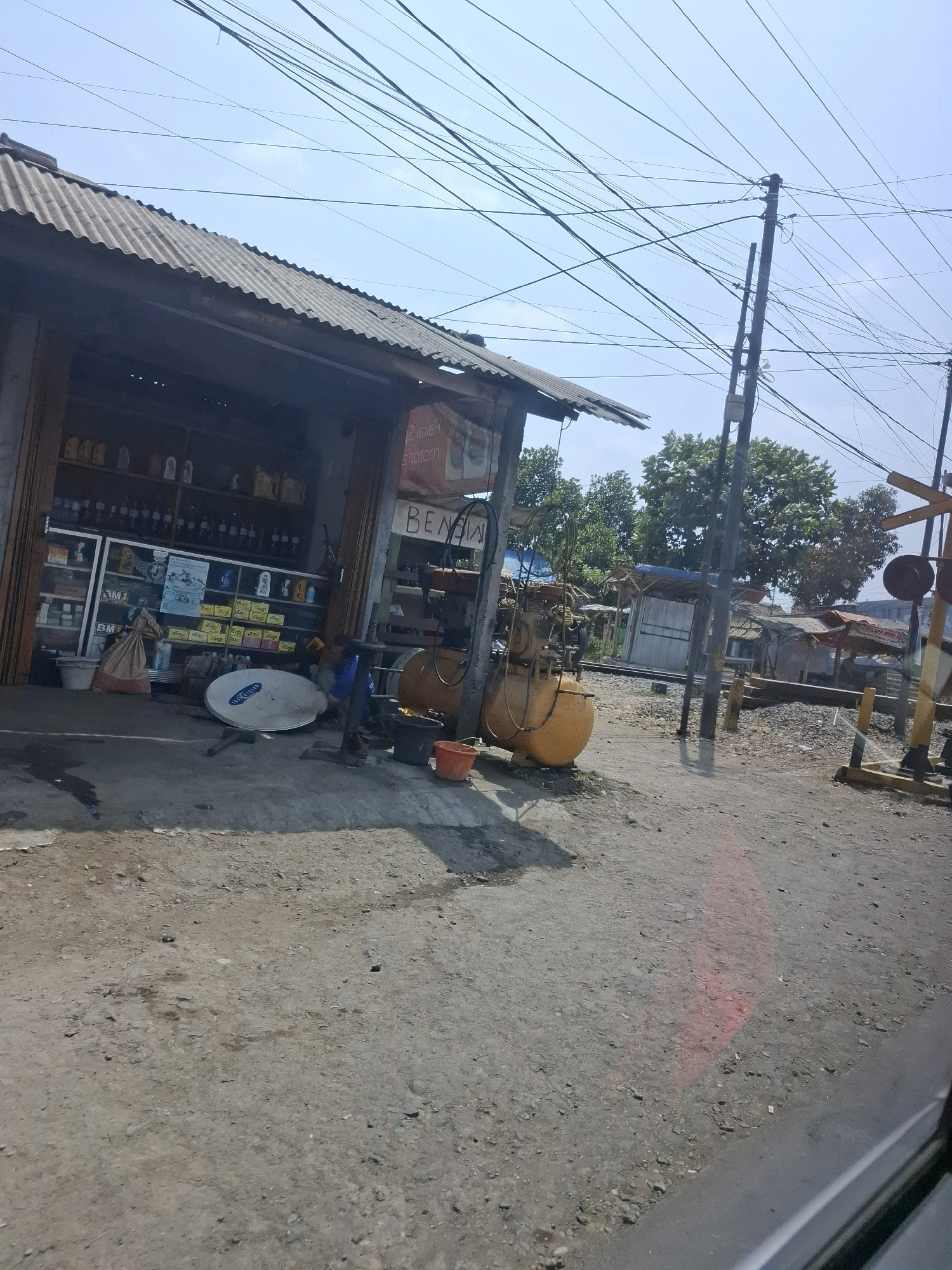 The image depicts a street scene in Lembursawah, Indonesia, likely in front of a small shop. The shop, with a corrugated metal roof and wooden framing, displays various goods on shelves, including bottles of oil and other automotive fluids. In front of the shop, a satellite dish rests on the ground, along with buckets and what appears to be an air compressor. A busy network of overhead electrical wires dominates the sky. To the right, a railway crossing with a signal post is visible. The ground is a mixture of dirt and gravel. The overall impression is of a functional, perhaps somewhat cluttered, area typical of a local business in a semi-urban or rural setting.
