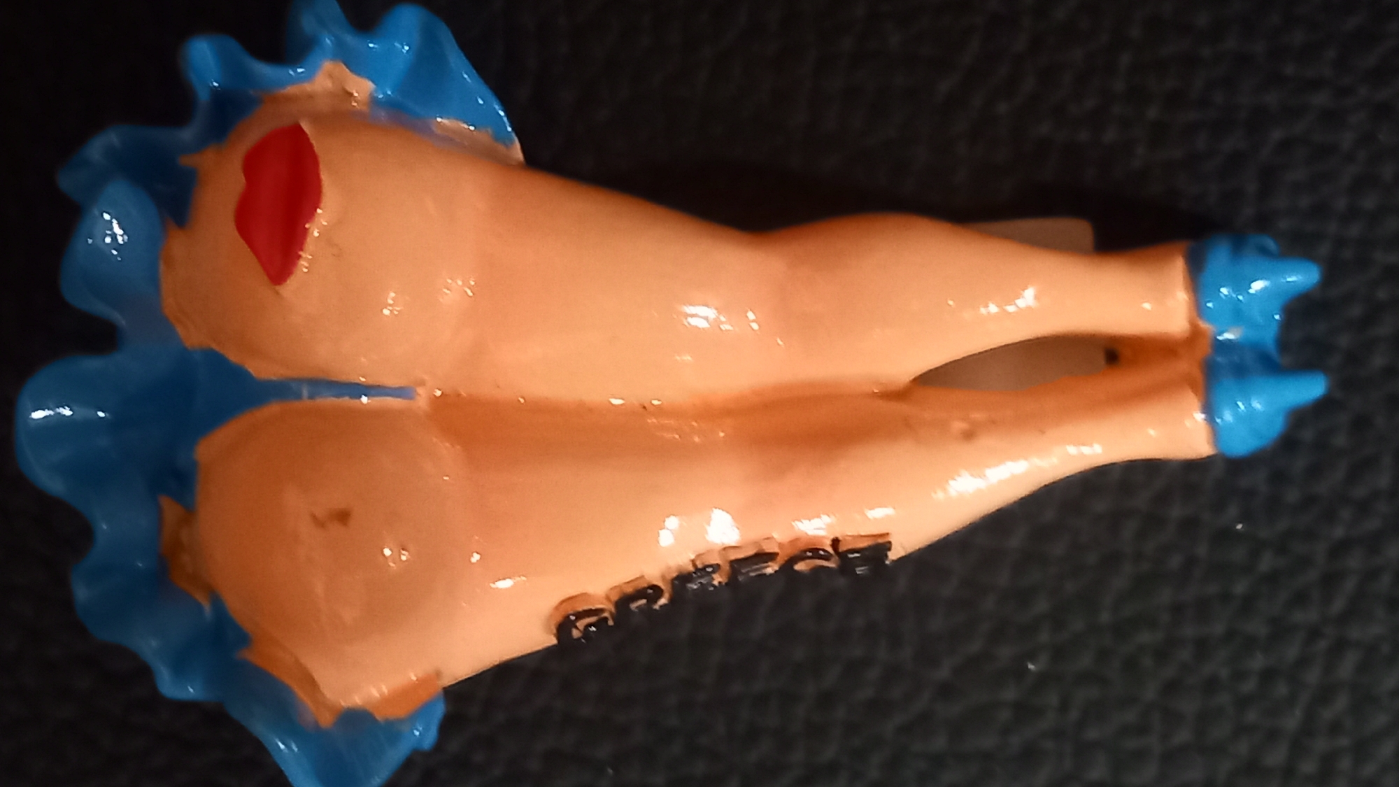 The image is a close-up, top-down view of a novelty item shaped like a pair of bare legs with blue high heels and a blue ruffled trim. The legs are a peachy-pink color and have red lipstick applied to the buttocks area, resembling a face. Black text, possibly a brand name or inscription, is visible running vertically down the left leg. The item is placed on a dark, textured surface, likely fabric. The overall impression is that of a kitschy souvenir or decorative object. No specific location in the city of Kos, Greece, can be determined from the image alone.