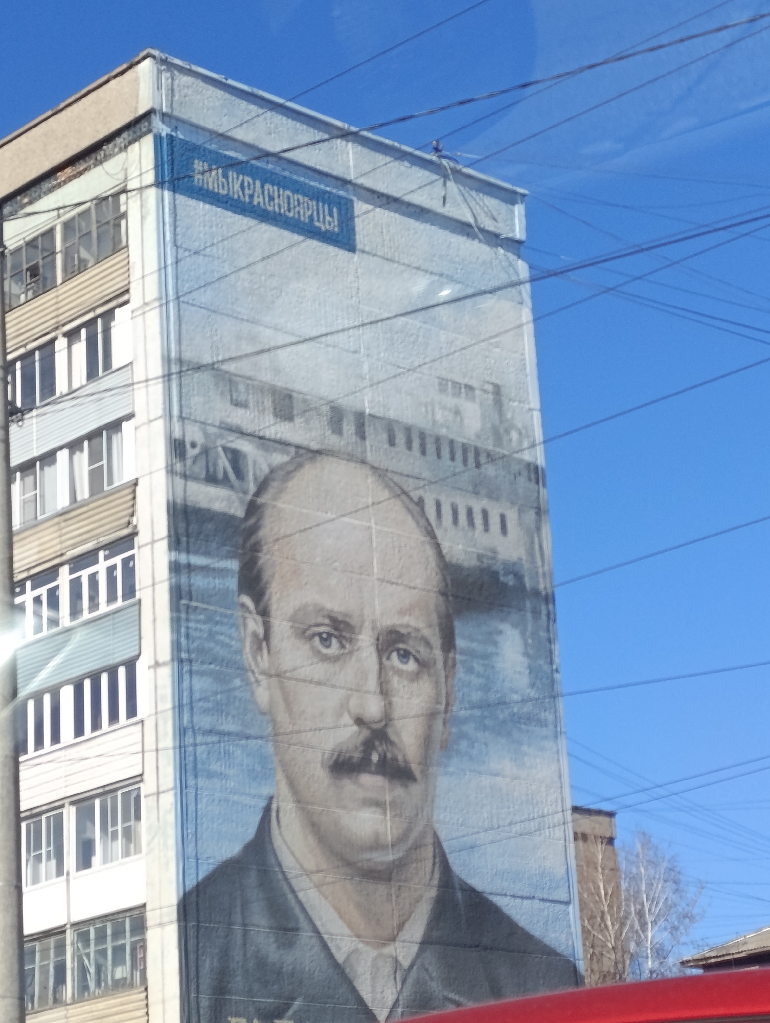 A tall, light grey building in Krasnoyarsk, Russia, features a large mural on its side. The mural depicts a man with a prominent mustache, wearing a dark suit. Behind the man in the mural is a structure resembling a bridge or industrial building over a body of water. At the top of the mural, a blue banner displays the text "#МЫКРАСНОЯРЦЫ". The building is positioned against a bright blue sky with several power lines crossing the view. Parts of other buildings and a tree are visible in the background. A portion of a red vehicle is seen in the lower foreground.