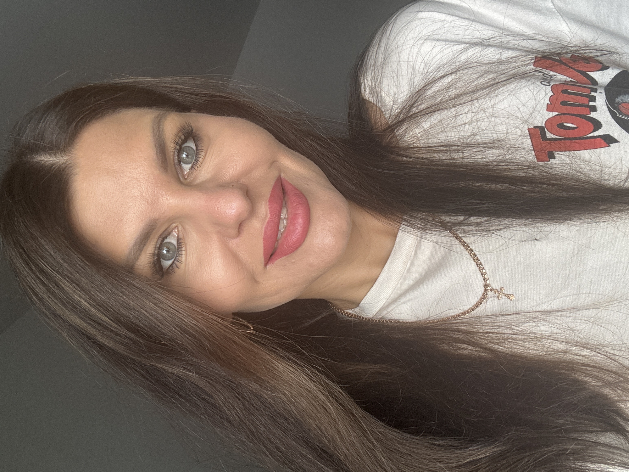 A medium shot captures a woman with long, dark brown hair, styled with a center part and falling loosely around her face and shoulders. She has fair skin, light blue-green eyes with noticeable eyelashes, and is wearing bright pink lipstick. Her attire consists of a white t-shirt with red lettering that reads "Tom and Jerry" and a graphic of a record. A delicate rose gold chain with a small cross pendant rests on her neckline. The background is a plain, dark gray wall, suggesting an indoor setting. The lighting appears natural, casting soft shadows on her face and highlighting her features. There is no indication of the city of Chaikovsky, Russia within the image.