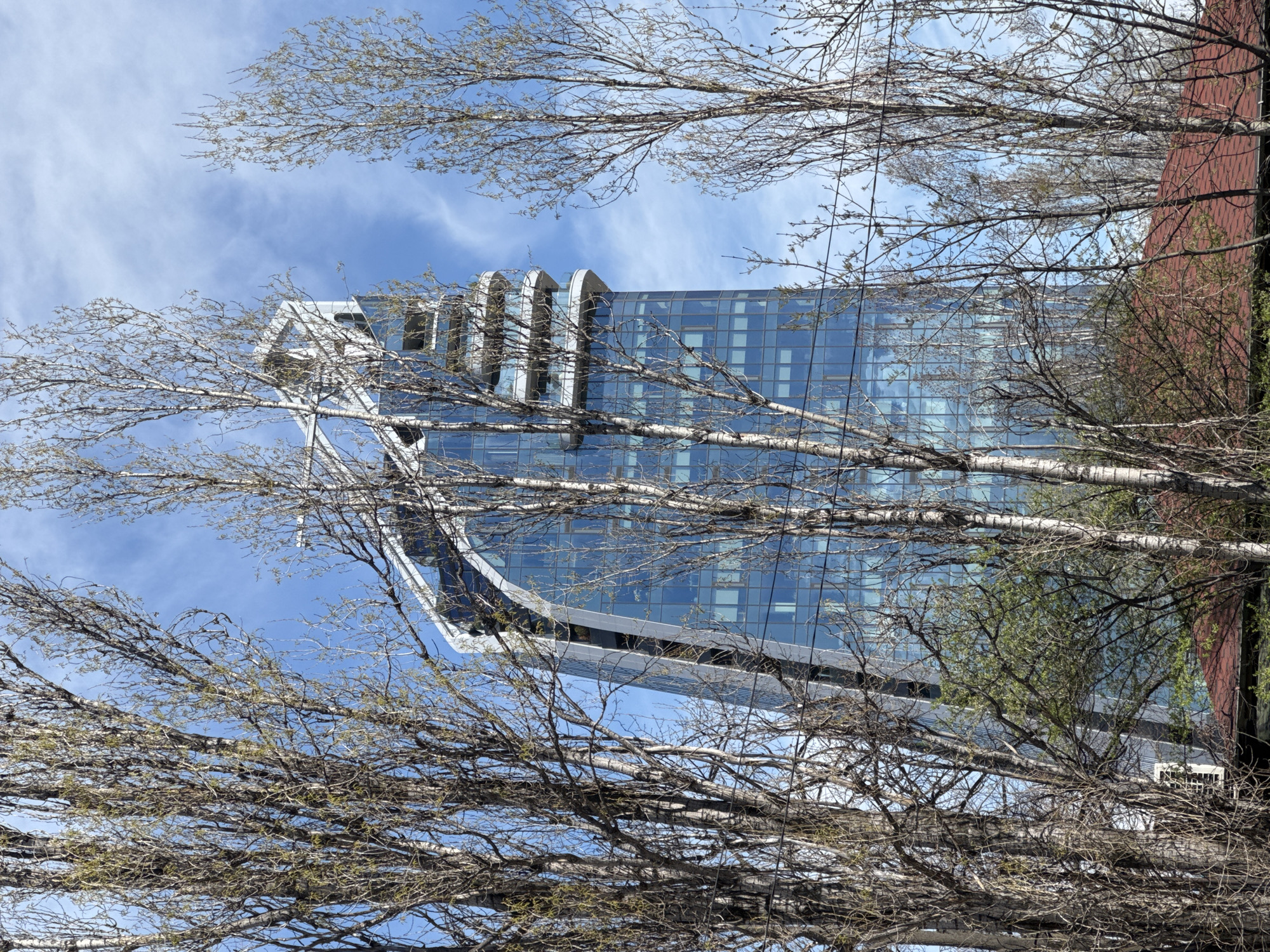 A low-angle view of a tall, modern building in Sofia, Bulgaria, taken during the day with trees in the foreground. The building has a blue glass facade and a unique architectural design with rounded elements at the top. The sky is partly cloudy with a blue background. The trees in the foreground have bare branches with some budding leaves, indicating early spring. A red brick building is partially visible to the right of the glass tower.