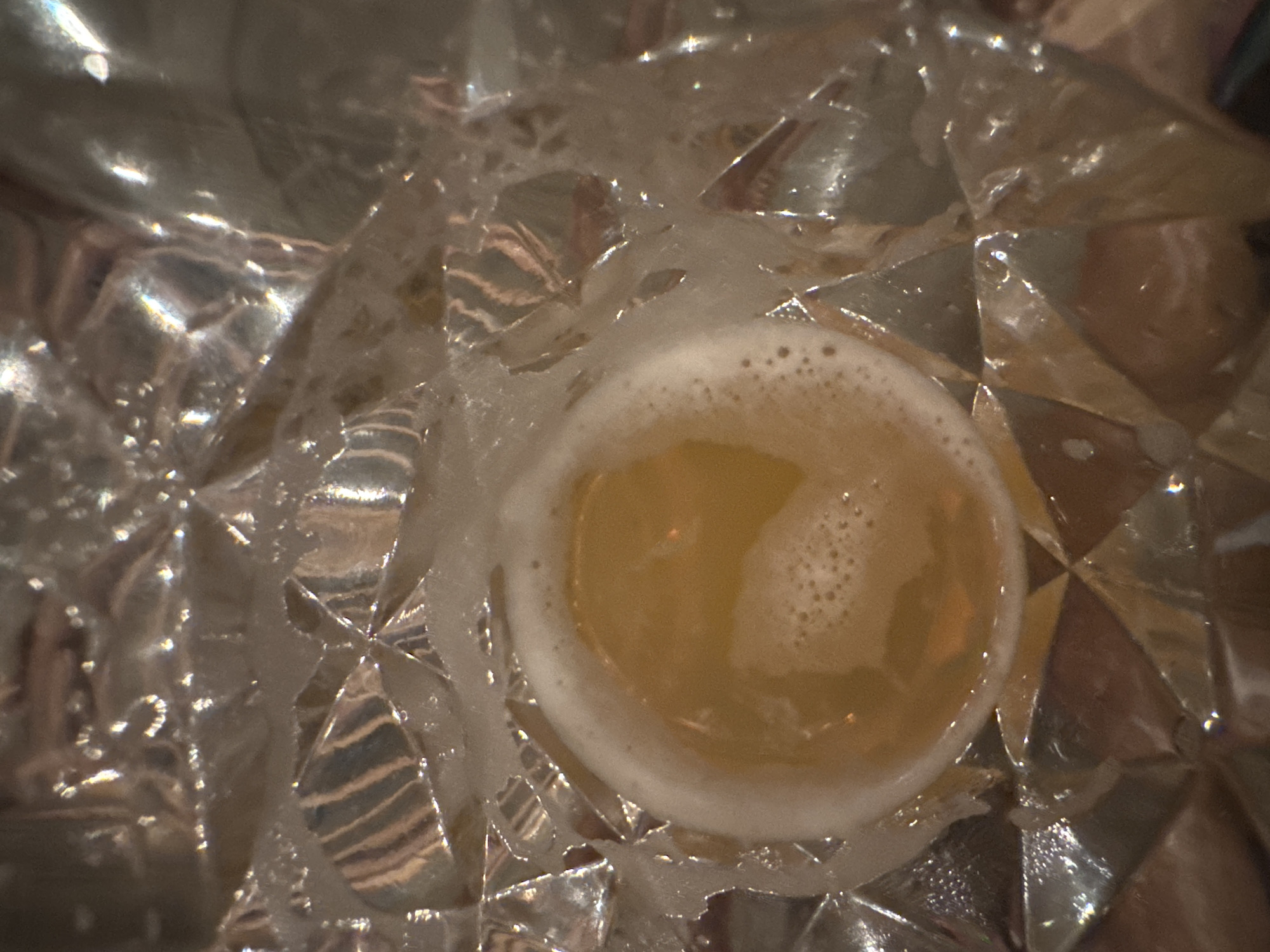 A close-up, overhead shot shows a glass of amber-colored liquid with a foamy head, resting on a textured, reflective surface. The surface appears to be made of crystal or glass with a diamond-like pattern. The liquid is a warm, golden hue and the foam is white with small bubbles. The lighting creates highlights and shadows across the scene, emphasizing the reflective qualities of the glass and the texture of the surface. No people or specific environmental details of Chuguyevka, Russia are visible in this image.