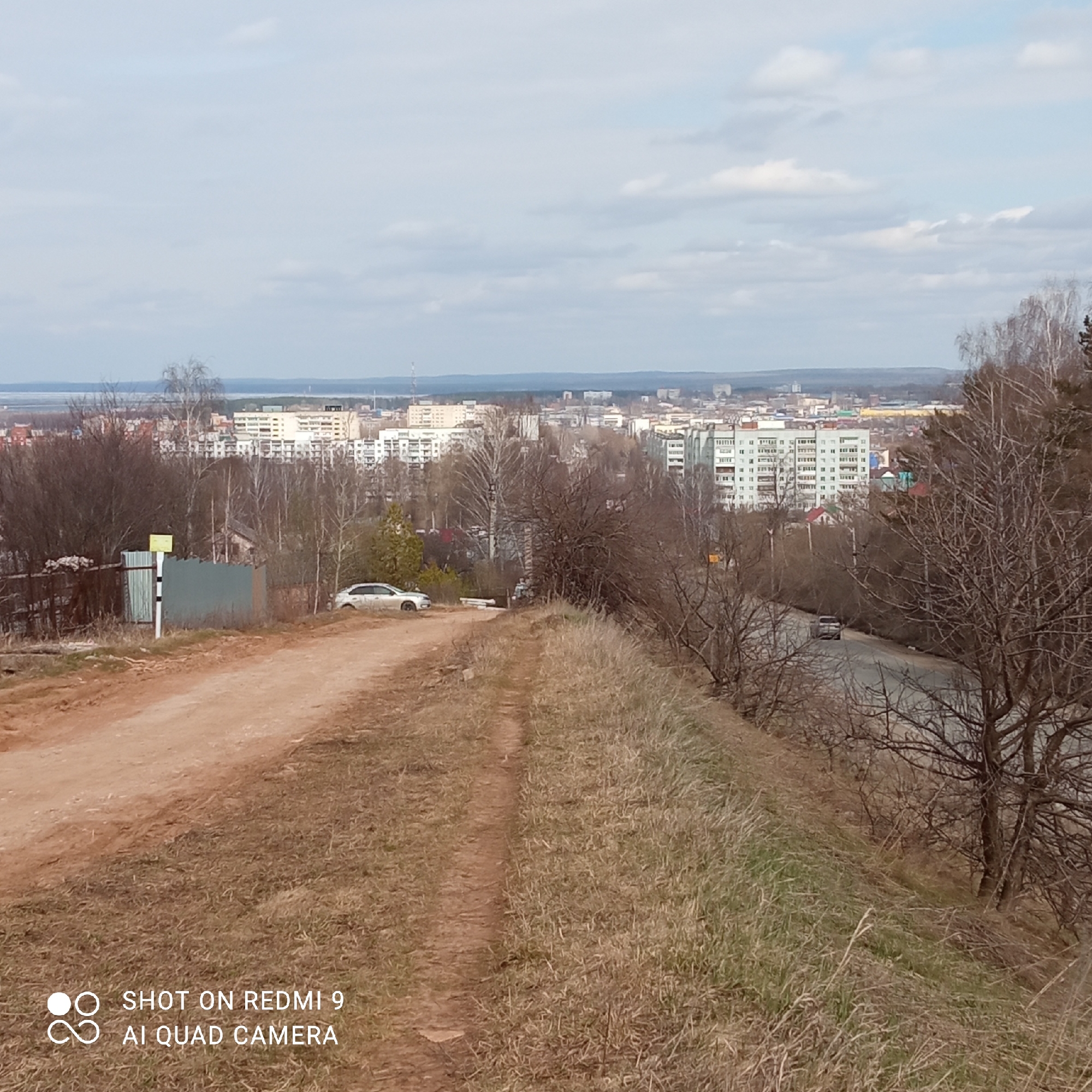The image shows a dirt road leading from an elevated position overlooking a cityscape. In the distance, buildings are visible, including residential blocks. A body of water, possibly a reservoir, borders the edge of the city. Adjacent to the road, there are patches of dry grass. A car is parked near the road. A road sign is also next to the road. The location is most likely Chaikovsky, Russia. A road runs behind and beside the road.