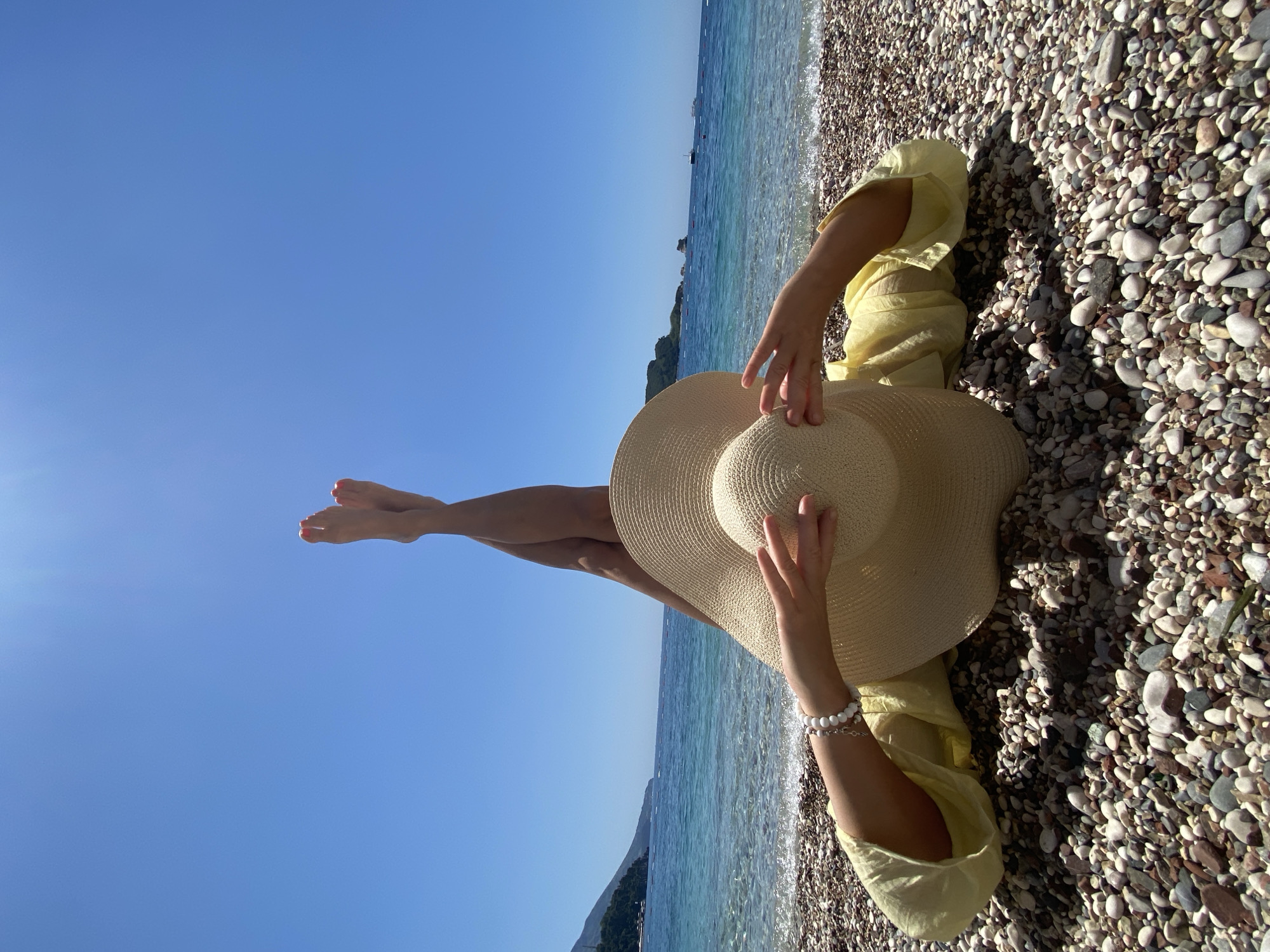 An individual is depicted lying prone on a multi-colored pebble beach. Their legs are raised vertically, crossed at the ankles, with feet showing red-painted toenails against a clear, bright blue sky. Their face is obscured by a wide-brimmed, light-colored straw hat, held in place by their hands; the left wrist is adorned with a white beaded bracelet. They are wearing a loose-fitting, light yellow long-sleeved top. The background features calm, clear turquoise water of the Adriatic Sea, gently washing ashore. In the distance, a vegetated mountainous coastline is visible on the left, while several rocky islets and scattered boats are present further out in the sea. The scene is located on a beach within the Budva Riviera, Montenegro.