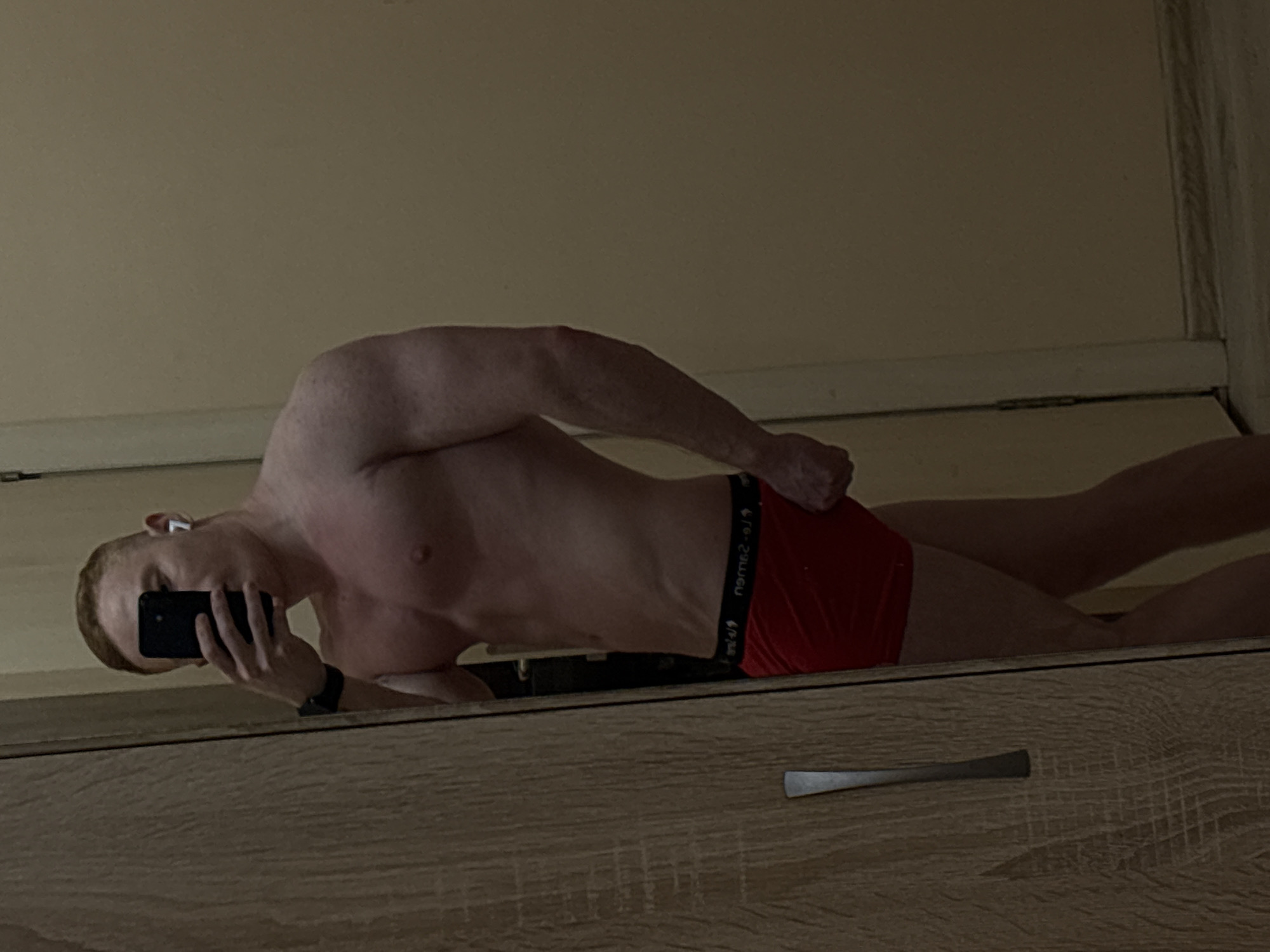 A person, appearing to be male, is captured in a selfie taken in Chaikovsky, Russia. The individual is shirtless and wearing red boxer briefs, lying on their side on a reflective surface, which is likely the top of a wooden dresser or cabinet. The image is a mirror reflection, causing the text and the person's posture to appear reversed from a direct view.
The subject has fair skin and light-colored hair. They are holding a black smartphone with their right hand to take the photo, largely obscuring their face. A white earbud is visible in their left ear, and a dark-colored wrist watch is worn on their left wrist. The pose is casual and relaxed.
The background consists of a plain, light-colored wall. In the foreground of the reflection, the wooden surface on which the person is lying shows distinct grain patterns and features a metallic, elongated handle, indicating it is a piece of furniture.
Visible text on the waistband of the red boxer briefs, due to the mirror reflection, appears as "oJt-2-9วนดอ" and other indistinct characters, likely a reversed brand name. The overall lighting is somewhat dim, suggesting an indoor setting without bright direct light. The activity is self-portrait photography in a casual, relaxed pose within an indoor environment.