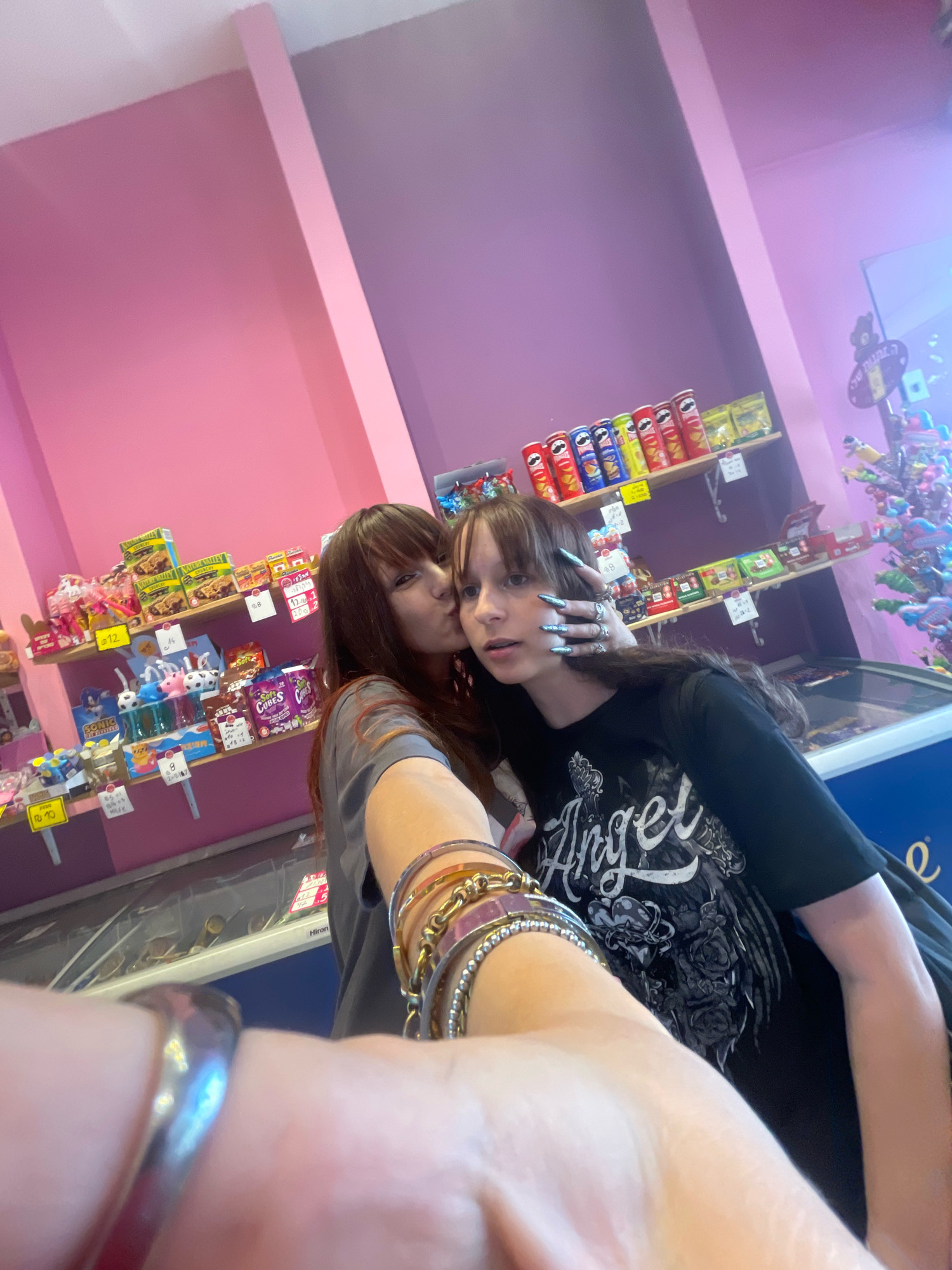 Two young females, one with long brown hair and bangs, is kissing the other on the cheek. They are in a shop with shelves stocked with candy and snacks. The walls are painted pink and purple. The person being kissed is wearing a black t-shirt with a graphic print. The person kissing has long, light-colored fingernails, and both are wearing bracelets. The scene is captured from a first-person perspective, as if taking a selfie. The presence of the shop and the specific products suggest a location within Raanana, Israel.