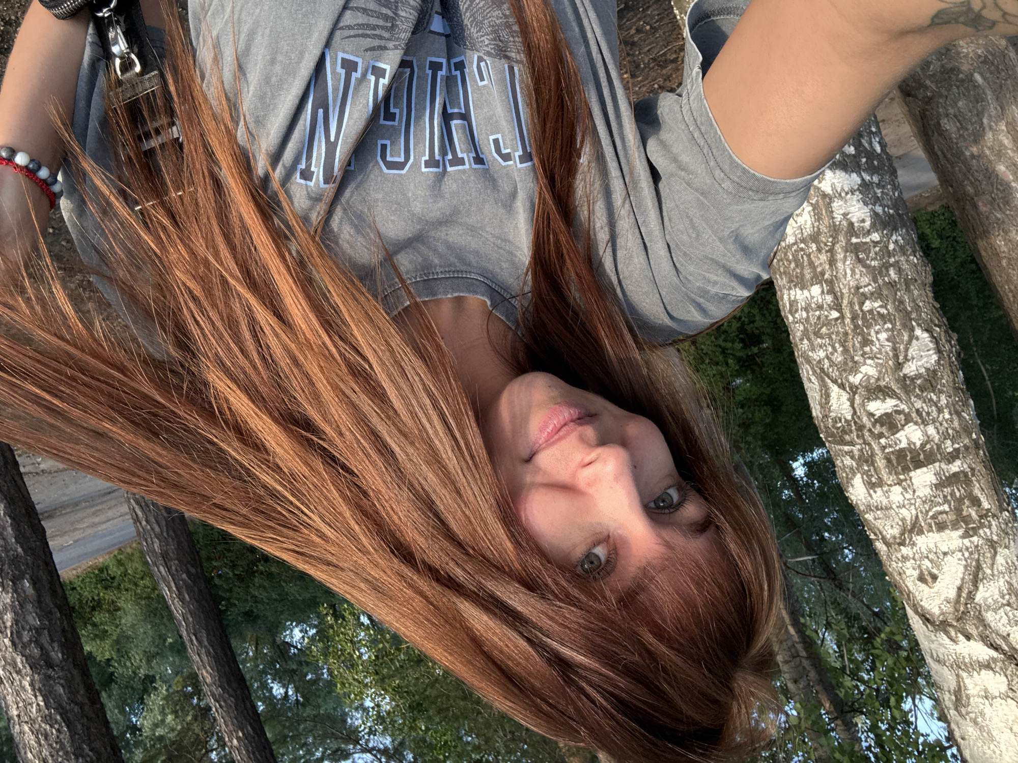 A young woman with long, auburn hair and green eyes is taking a selfie outdoors. She is wearing a grey t-shirt with blue lettering and a graphic design on it. The background consists of trees, including a birch tree to her left and pine trees to her right. A paved path is visible behind the pine trees, suggesting the location is a park or roadside. The lighting indicates it is likely late afternoon or early evening. The woman appears to be in a relaxed and natural setting. The city of Chaikovsky, Russia is known for its parks and natural scenery, making this location plausible.