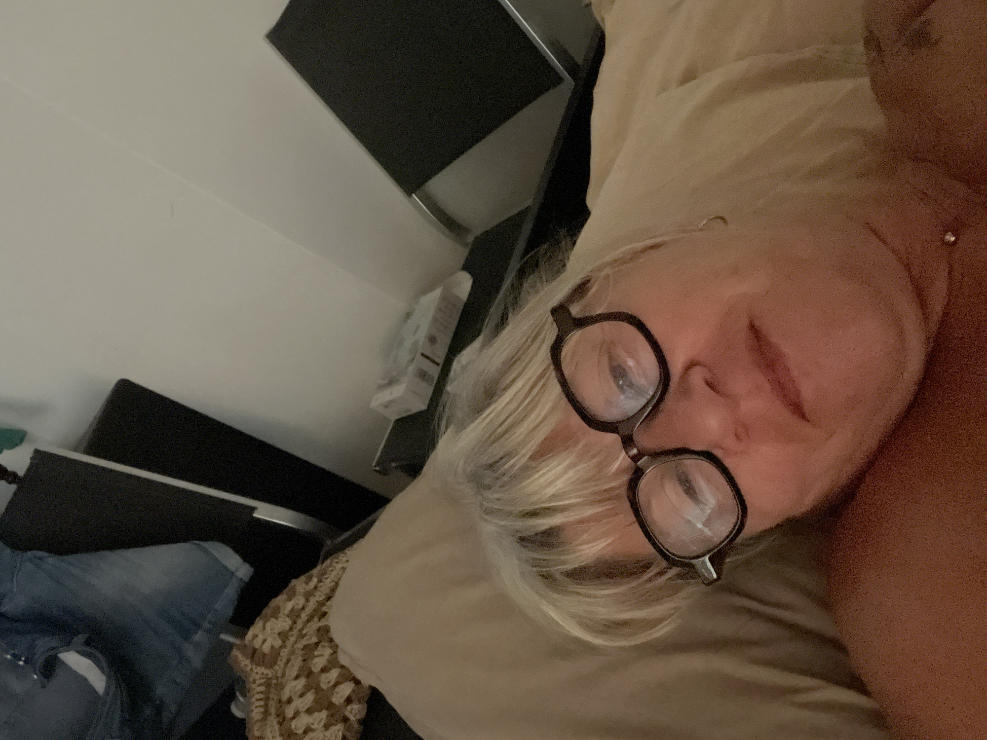 A woman with blonde hair and glasses is lying in bed. She is wearing a necklace. The background shows a bedside table with a blanket and some clothing. The lighting is dim. The image appears to be taken from a close-up perspective, with the woman's face filling most of the frame. The overall impression is one of casual intimacy, as if the photo was taken in a private setting.