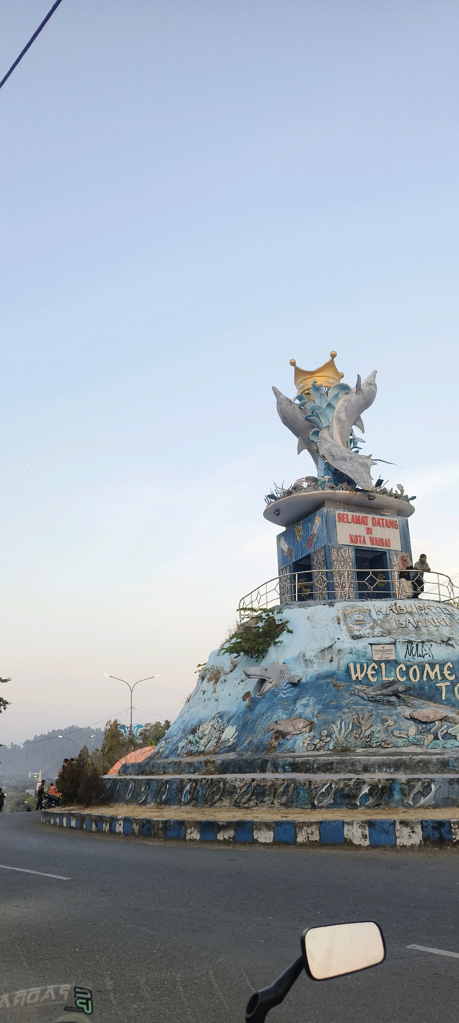 A monument with sculptures of dolphins and a crown is situated on a hillside next to a road in Waisai, Indonesia. The monument features a blue and white facade decorated with marine life motifs. The inscription on the monument reads "SELAMAT DATANG DI KOTA WAISAI" and "WELCOME TO". Two individuals are visible standing on a platform of the monument. The sky is clear and light blue, suggesting it is daytime. The surrounding environment includes a road, trees, and hills in the background.