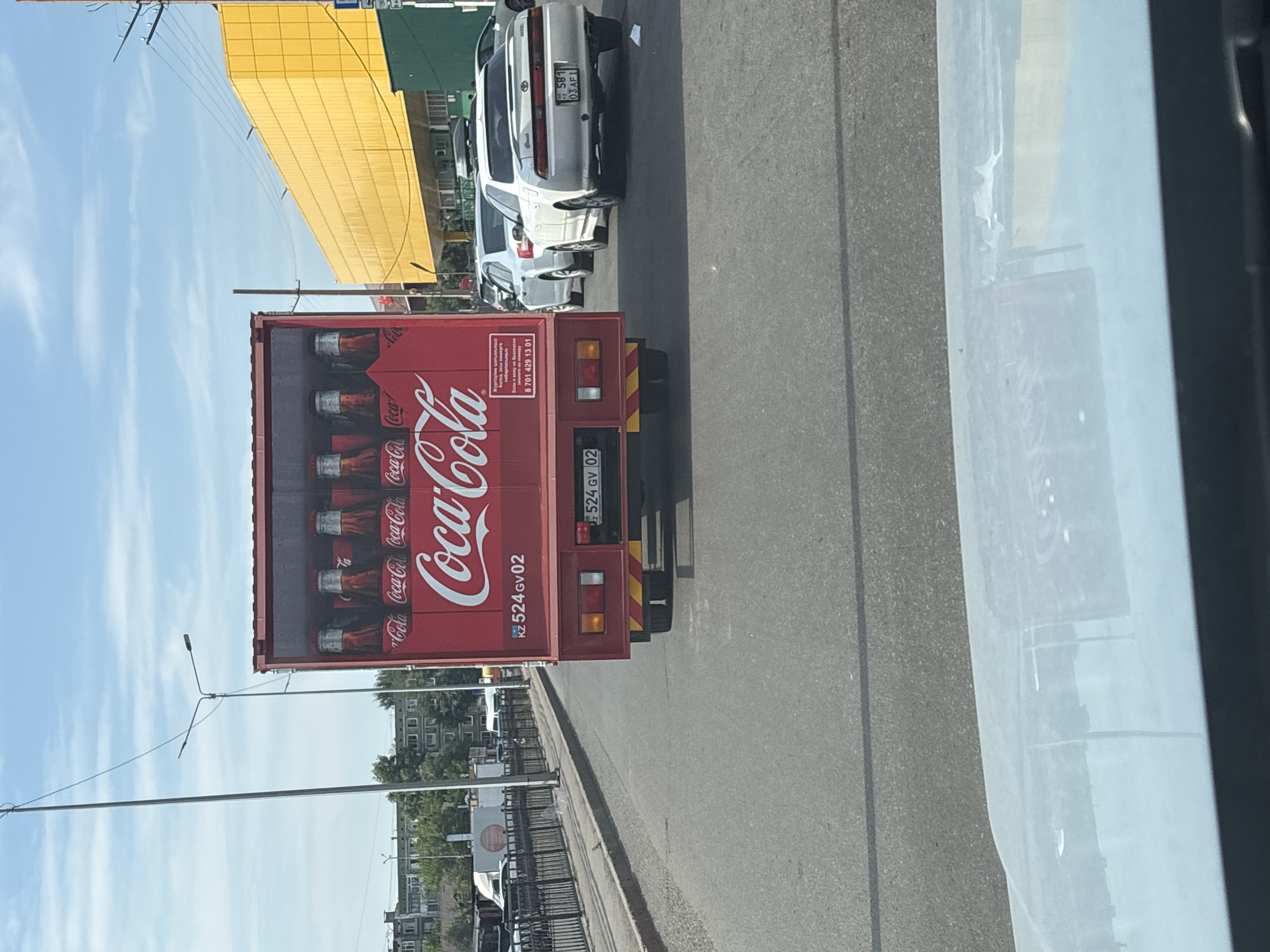 The image captures a daytime scene on a street in Astana, Kazakhstan. The primary focus is a red Coca-Cola branded truck. The side of the truck prominently displays the Coca-Cola logo and images of Coca-Cola bottles. Text on the truck includes "Coca-Cola" repeated multiple times, a license plate reading "KZ 524GV02", and some text in what appears to be Kazakh and Russian, along with a phone number "8 701 429 13 01".
Behind the truck, several white cars are parked or driving on the road, with one car's license plate visible as "581 03 AF1". In the background, a large, bright yellow building stands out, suggesting an urban or commercial area. A green structure is also visible next to the yellow building. The sky is blue with some white clouds, indicating clear weather. The scene appears to be captured from another vehicle, possibly indicating traffic or movement. The overall impression is a snapshot of daily life in a city, with a focus on a commercial vehicle.