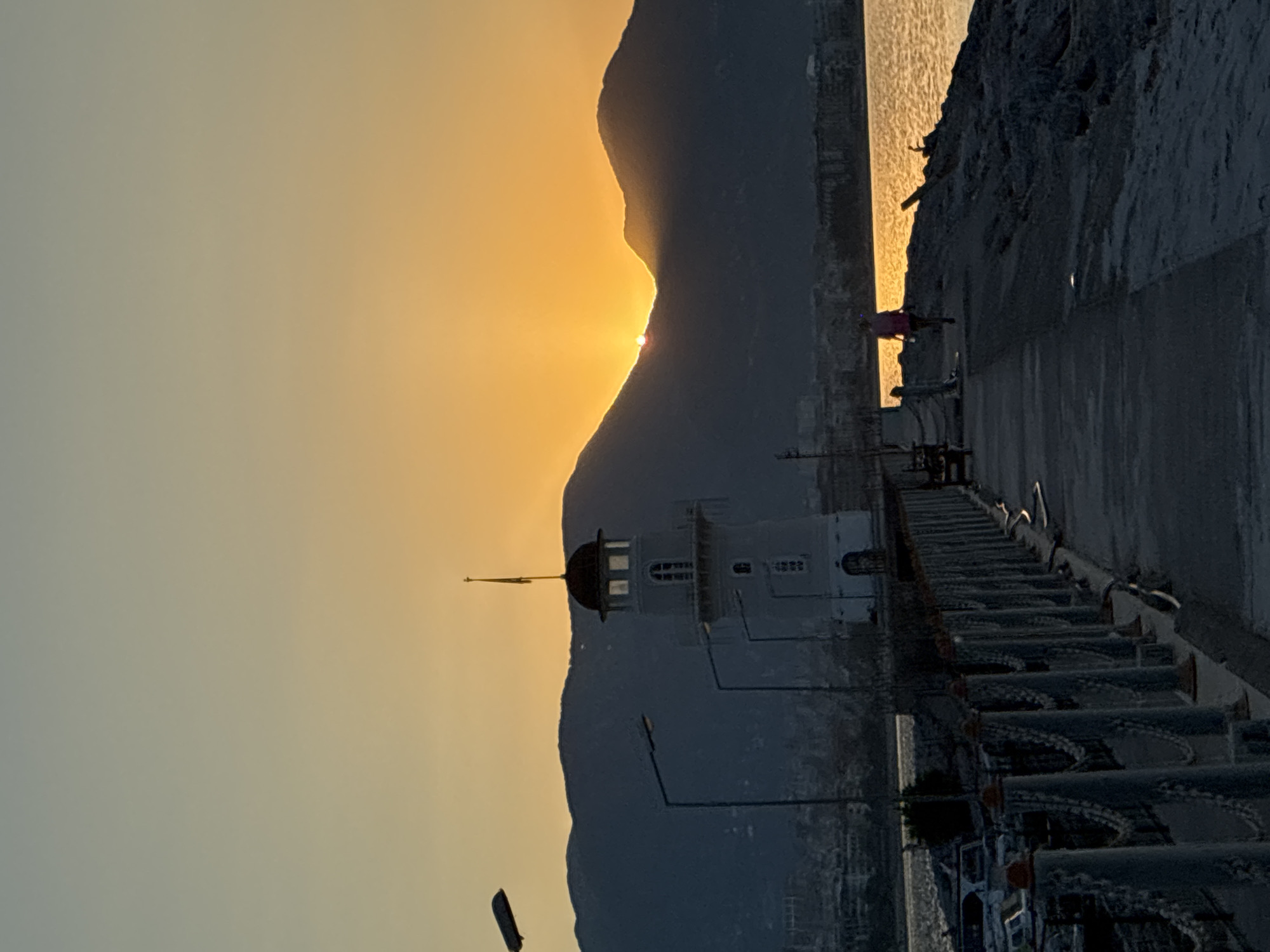 The image captures a scenic view of Alanya, Turkey, during sunset. The foreground shows a paved walkway leading to a stone-built pier, lined with bollards and chains. To the left, a lighthouse stands prominently, its white structure contrasting with the darkening sky. Several lampposts are visible along the walkway. In the mid-ground, a person is seen walking along the pier, silhouetted against the warm glow of the setting sun. The background features a range of mountains, partially obscuring the distant city skyline. The sun is setting behind the mountains, casting a golden hue over the water and the sky.