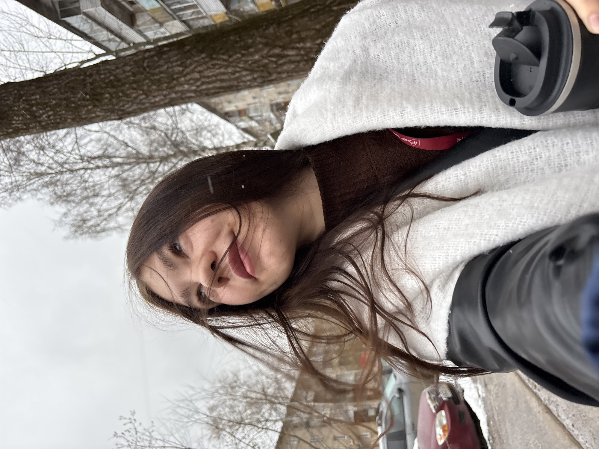 The image shows a woman in Perm, Russia, in an outdoor setting on a cold, likely winter day, indicated by bare trees and overcast sky. She is wearing a brown top, black jacket, and a white, textured wrap. She is holding a black travel mug. The backdrop consists of a tree, a building with visible windows, and parked cars along a street.