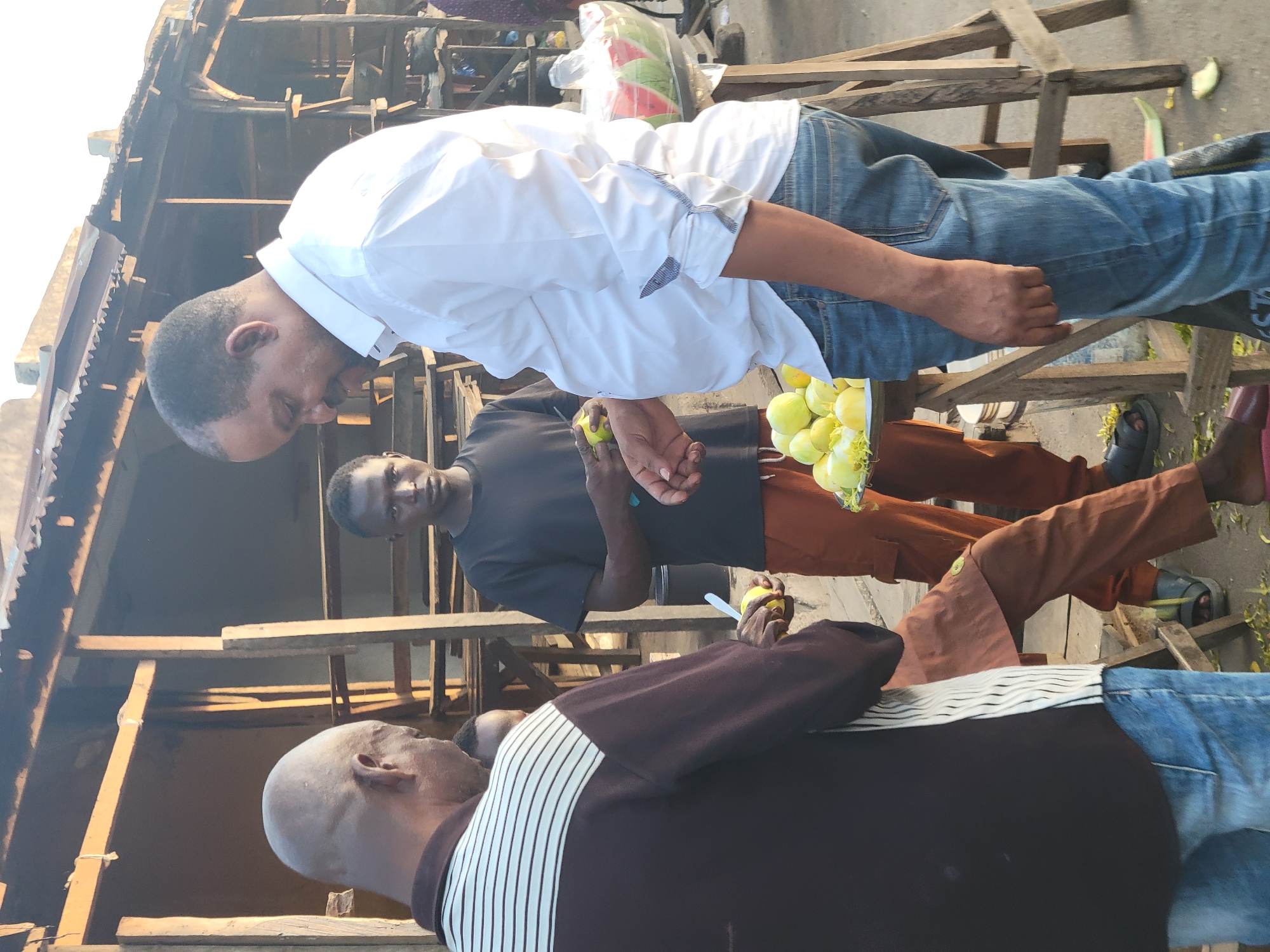 Three men are standing near a wooden structure in Madalla, Nigeria. The man closest to the viewer has a shaved head and wears a dark brown shirt with white stripes, along with blue jeans. Another man wears a black shirt and brown trousers, holding a green fruit. A third man wears a white shirt and blue jeans. The location shows signs of an outdoor market or public area. A bag of watermelon is visible near the top of the image.