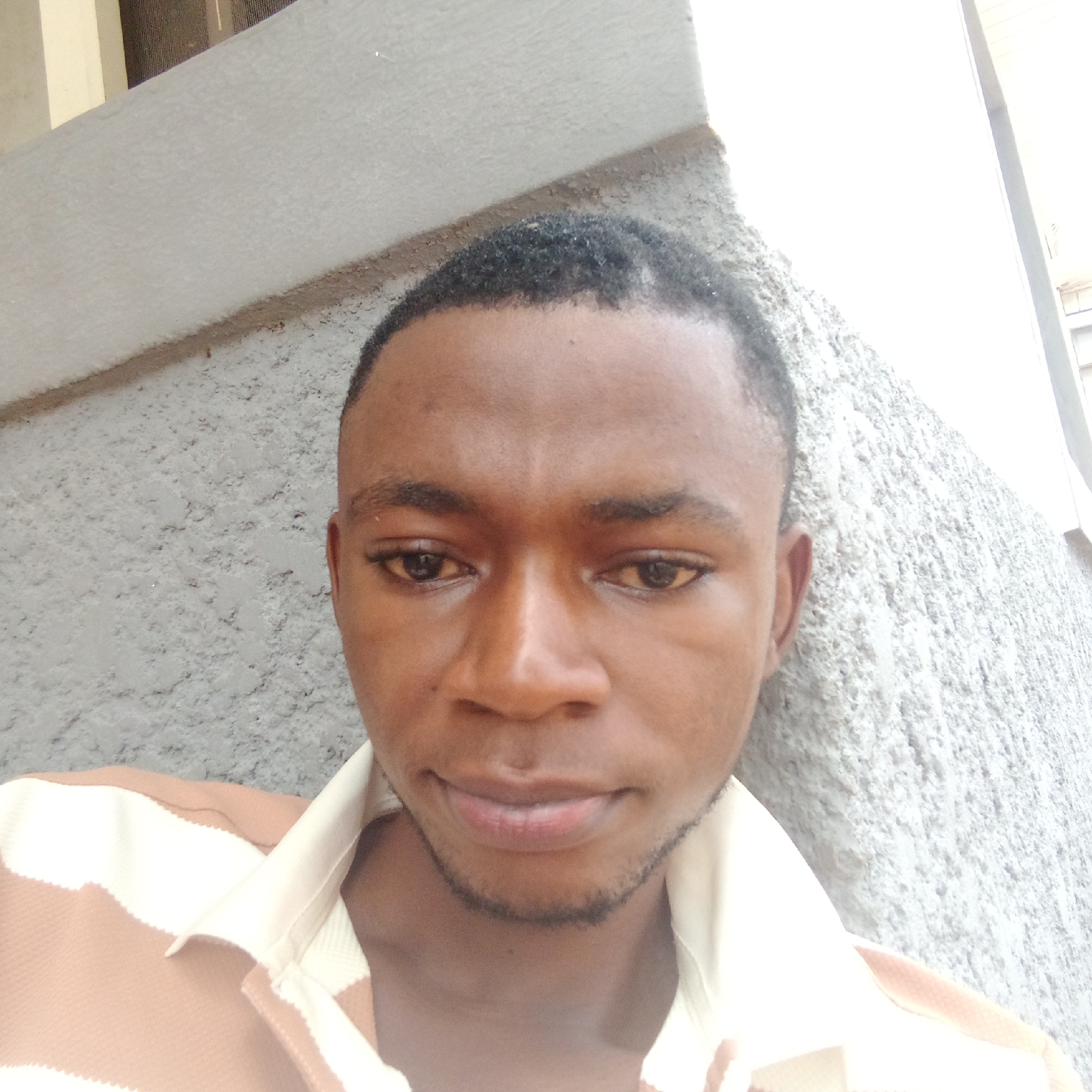 A young Black man with short black hair and a light beard is pictured in a close-up selfie. He is wearing a collared shirt with horizontal stripes of tan and white. The background appears to be an outdoor setting with a textured gray wall and a lighter colored structure to the left. The lighting suggests it is daytime. The man's gaze is directed towards the camera.