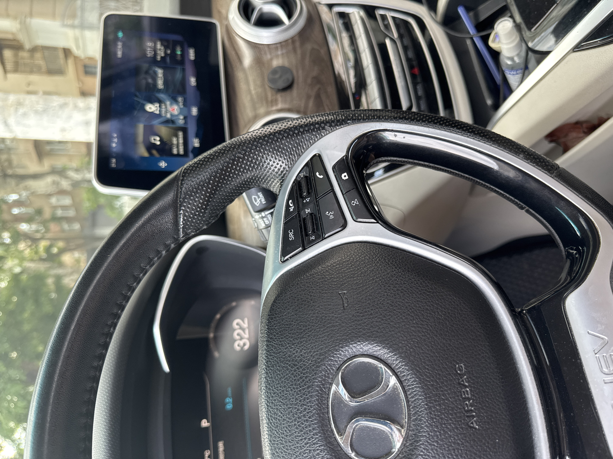 The image shows the interior of a car, specifically the steering wheel, dashboard, and infotainment screen. The steering wheel is black leather with silver accents and controls. The dashboard has a digital display showing "322" and other indicators. The infotainment screen displays a car's interface with various icons and text in what appears to be Chinese. The car's interior features wood-grain trim and circular air vents. A small bottle of hand sanitizer and a blue pen are visible in the center console area. The background suggests an urban environment with buildings and greenery, consistent with a city setting. The image does not provide any specific visual cues to confirm the location as Odesa, Ukraine.