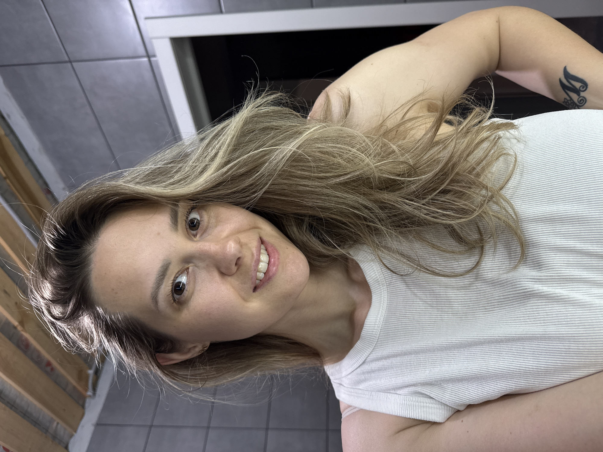 A woman with blonde hair smiles at the camera. She is wearing a white ribbed shirt. A tattoo is visible on her upper arm. The background includes grey tiles and a white door frame. The image does not contain enough information to confirm it was taken in Chaikovsky, Russia.