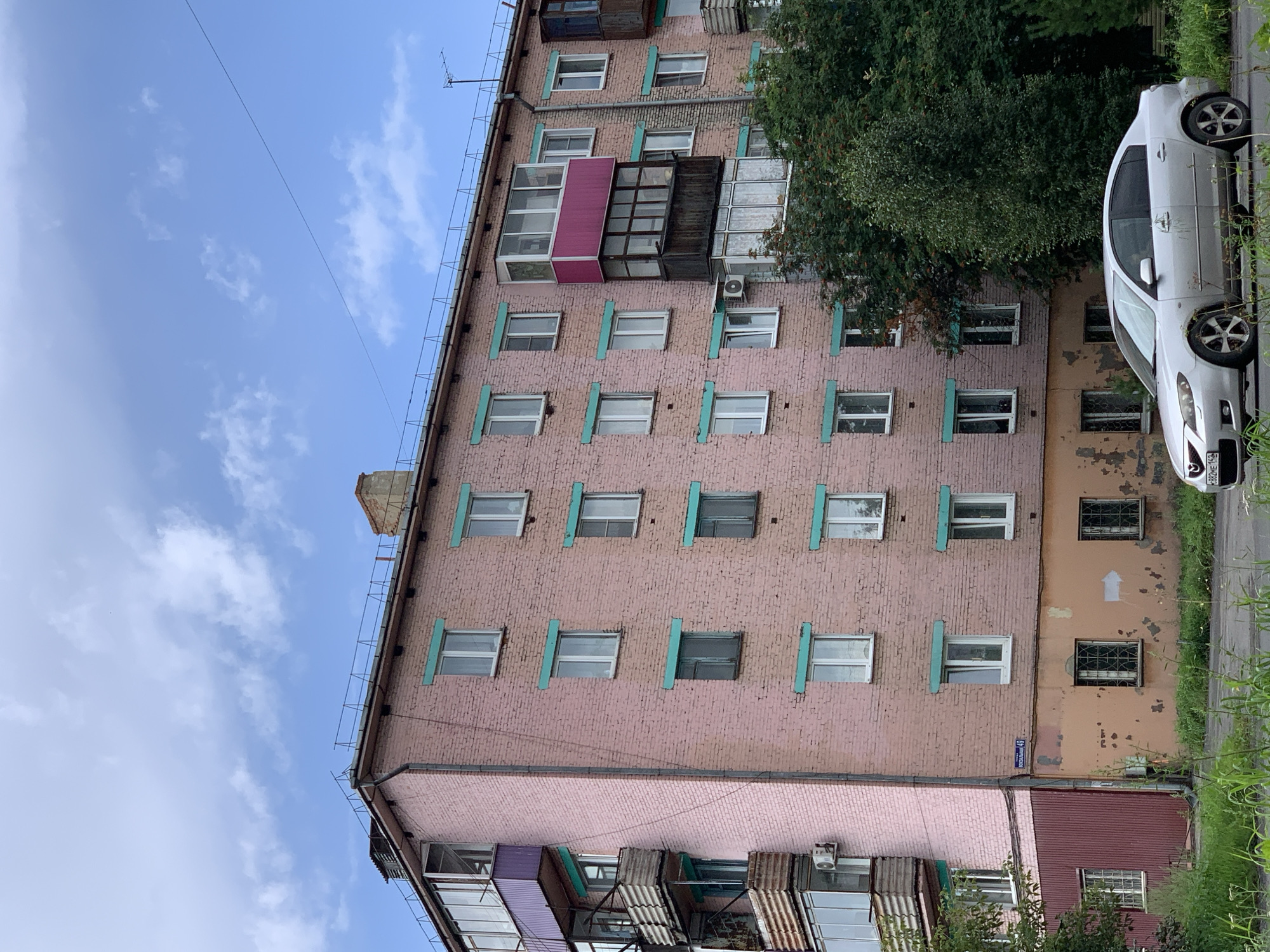 The image shows a pink brick apartment building in the city of Prokopyevsk, Russia. The building has multiple floors with numerous windows, some with teal-colored trim. Several balconies are visible, some enclosed and others open with weathered railings. A silver sedan is parked in front of the building on a paved road. Green grass and trees are present in the foreground and around the building. The sky is blue with scattered white clouds. A street sign with Cyrillic text is attached to the side of the building.