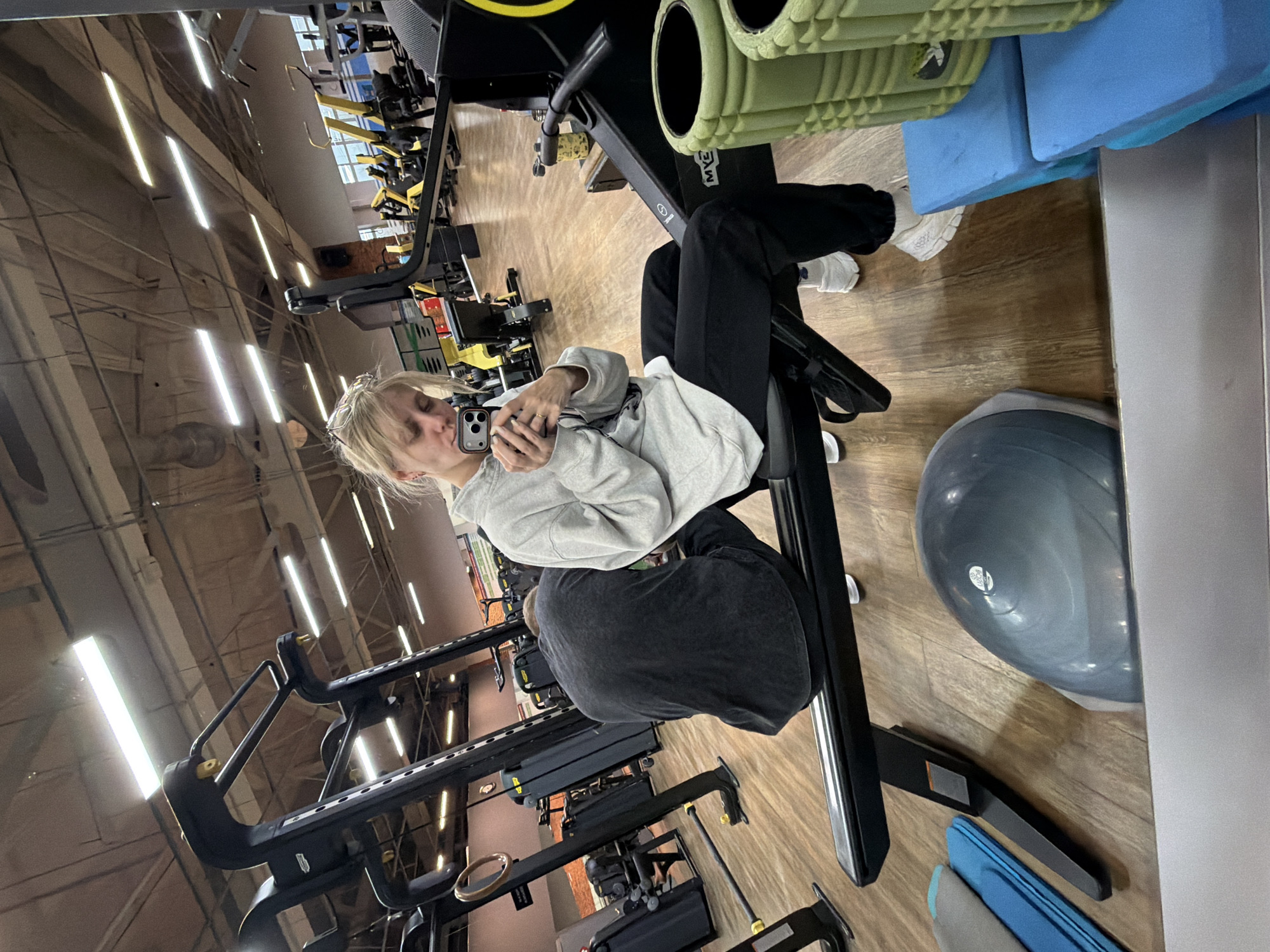 The image shows a woman holding a phone in a gym. The location is Chaikovsky, Russia. The woman is wearing a grey sweatshirt and black pants with white sneakers. She is sitting on a black exercise machine. There is a grey bosu ball on the floor, and green foam rollers and blue mats are stacked near the machine. Other gym equipment is visible in the background. The gym is well-lit.