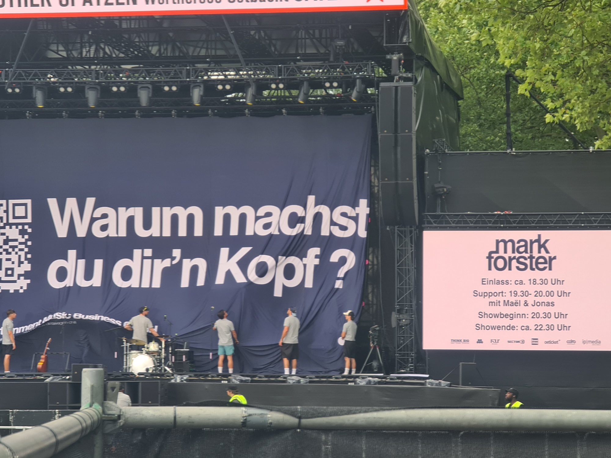 The image shows a stage setup for a concert in Klagenfurt, Austria, likely at the Wörthersee Stadium. A large dark blue banner dominates the center of the stage with large white German text that reads "Warum machst du dir'n Kopf?". To the left of the banner, a QR code is visible. On the stage, there are several people in casual attire, appearing to be part of the event crew or band members. One person is standing by a guitar. Another is seated at a drum set, and two others are standing near the banner. To the right of the stage, a large pink screen displays event information, including "mark forster", the artist's name, and show times. The screen also features logos of sponsors or partners. The background shows trees and the sky, indicating an outdoor venue. The overall scene suggests preparation for a musical performance.
