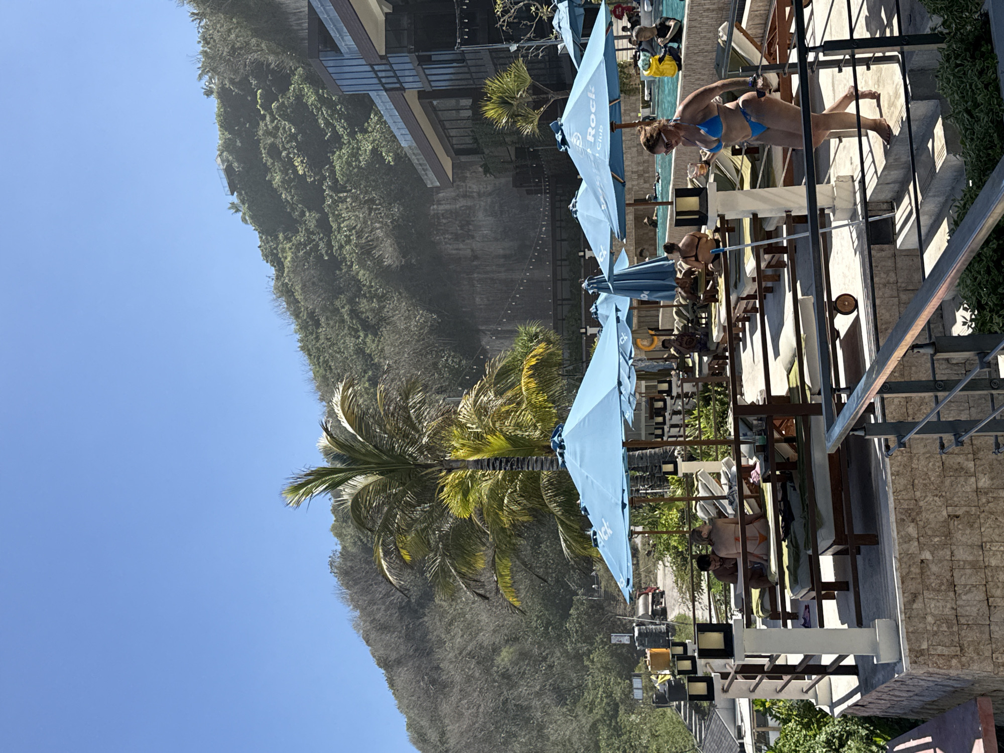An outdoor swimming pool area is depicted, with a steep, vegetated cliff face in the background. The cliff is covered in a mix of trees and dry brush, with a modern building constructed on its upper slopes. Several people are visible, some relaxing on lounge chairs under large blue umbrellas, while others are in the pool. One woman in a blue bikini is walking down a set of stairs in the foreground, holding a drink. The environment appears to be a resort or hotel in Kuta Selatan, Indonesia, suggested by the tropical vegetation and architectural style. The sky is clear and bright blue.
