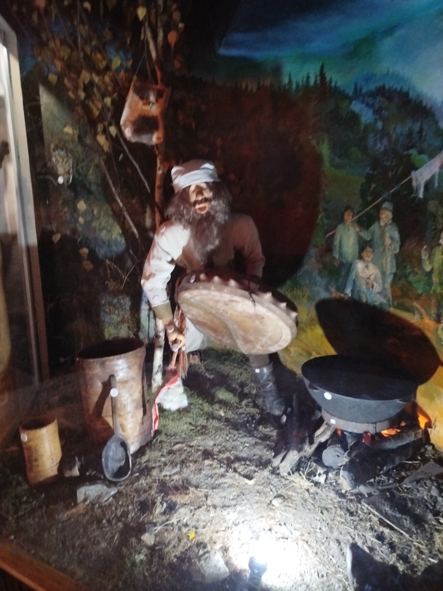 The image depicts a diorama in what appears to be a museum exhibit, possibly located in Tashtagol, Russia, given the context. The central figure is a man with a long, dark beard and a white head covering, dressed in traditional attire, holding a large drum. He is positioned in a rustic, outdoor setting with trees and foliage suggested in the background, which is painted with a nighttime forest scene. In front of the man, there is a fire pit with a large black pot or cauldron over it, and various wooden buckets and tools are scattered on the ground. The ground is covered with dirt, dry leaves, and some rocks. In the background, there are painted figures of other people in traditional clothing. The lighting in the scene is dramatic, highlighting the central figures and objects. The overall impression is a representation of historical or cultural life in a rural or wilderness environment.