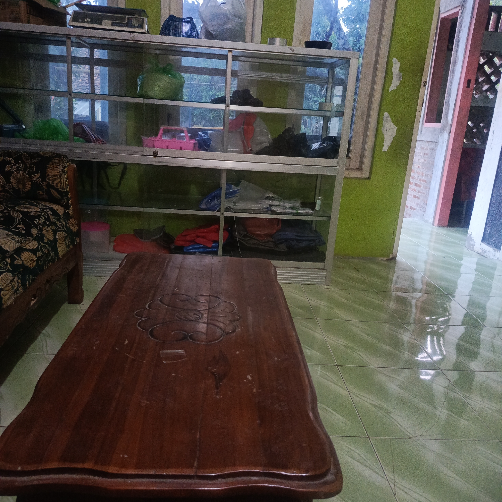 This is a medium shot, eye-level interior photograph taken in Karangtuang, Indonesia. The scene depicts a living space with a focus on a wooden coffee table in the foreground and a glass display cabinet in the background. The coffee table is dark brown, rectangular, and has carved decorative elements on its surface. Behind the table, a glass display cabinet with a silver frame stands against a lime-green wall. The cabinet contains various items, including plastic bags, clothing, and a pink toy car on the second shelf. To the left of the cabinet, a portion of a sofa with a dark patterned upholstery is visible. The floor is covered in light green square tiles that reflect the light. The background reveals a doorway and window, suggesting the presence of other rooms and the outdoors. The lighting is dim, indicating it may be evening or an overcast day. There are no people visible in the image, and no active interactions are occurring. The overall impression is that of a lived-in but somewhat cluttered domestic environment.