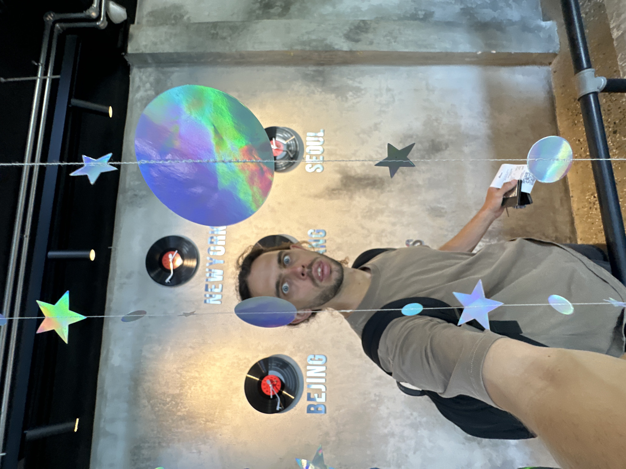A man with brown hair and a beard, wearing a tan t-shirt and black backpack, takes a selfie in front of a wall with circular clocks representing different cities and their names below in silver lettering. The cities visible are Beijing, New York, and Seoul. The wall is textured gray concrete, and the lighting is warm. Suspended from the ceiling are strings adorned with colorful holographic stars and discs. The man is holding a boarding pass in his right hand. The background shows a black ceiling with light fixtures and what appears to be a railing in the foreground. The scene appears to be in a modern, possibly airport-themed, environment. This location is likely within the city of Đà Nẵng, Vietnam, possibly at the Đà Nẵng International Airport or a related attraction.