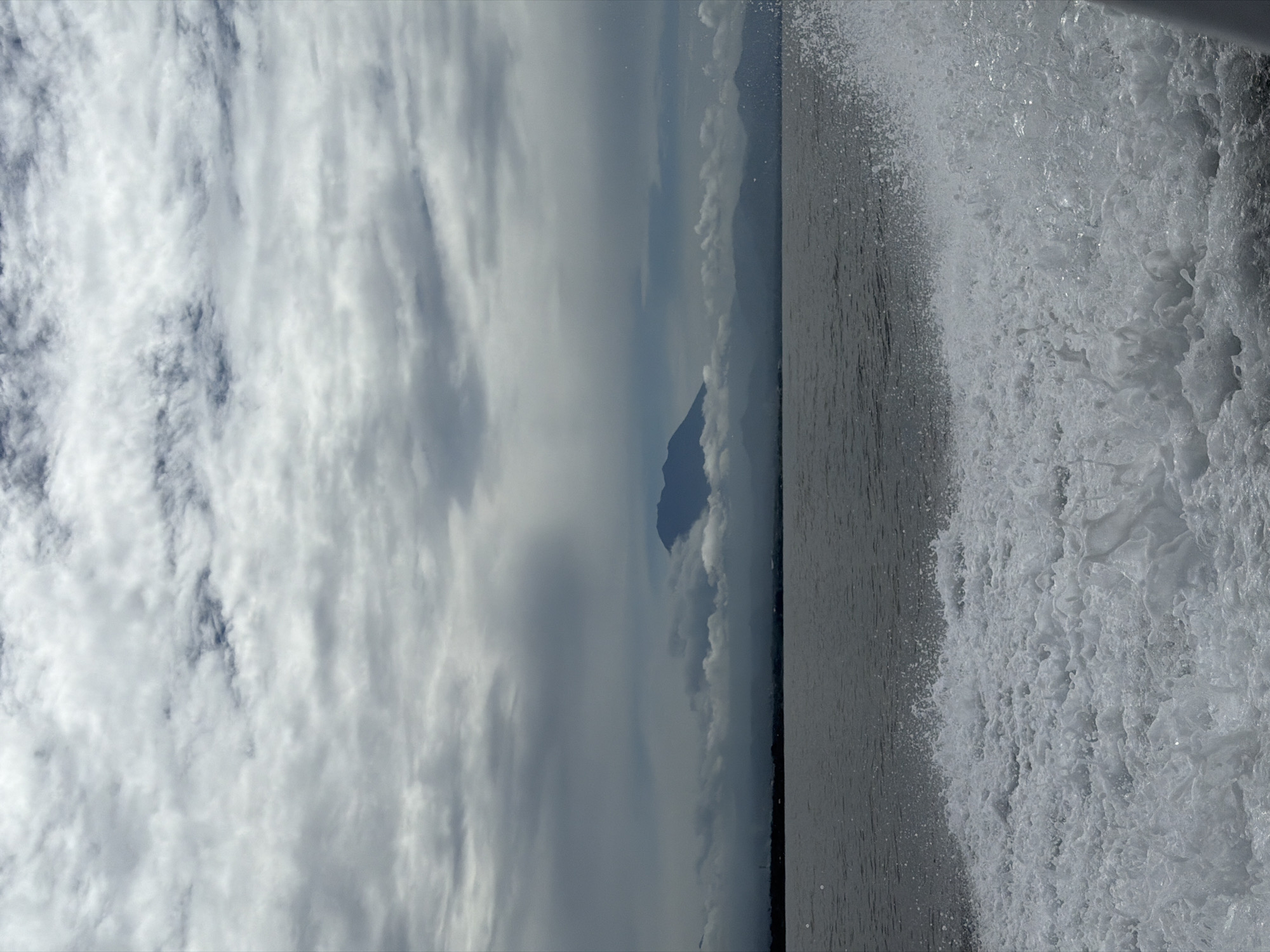 The image shows a landscape featuring a body of water, possibly a lake or sea, with waves visible on the right side of the frame. In the distance, a mountain silhouette is partially obscured by clouds. The sky is filled with a mix of clouds, some dense and others scattered, creating a cloudy atmosphere. The picture appears to be taken from the side of a moving vessel. Location: Unknown, Indonesia.