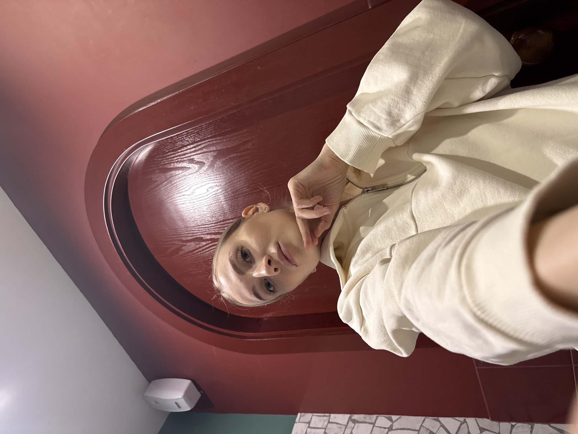 A person is posing for a selfie in what appears to be a bathroom. The person has fair skin, light blonde hair, and blue eyes. She is wearing a light-colored zip-up hoodie. She is holding her hand up to her face in a contemplative pose.
The background includes a dark red, rounded doorway or archway, and walls painted in a similar red hue. The bottom of the frame shows a glimpse of tiled flooring in a mosaic pattern. A white wall-mounted dispenser is also visible.
The photo may be taken in a commercial setting. The city of Izhevsk, Russia, is not discernible from the image.