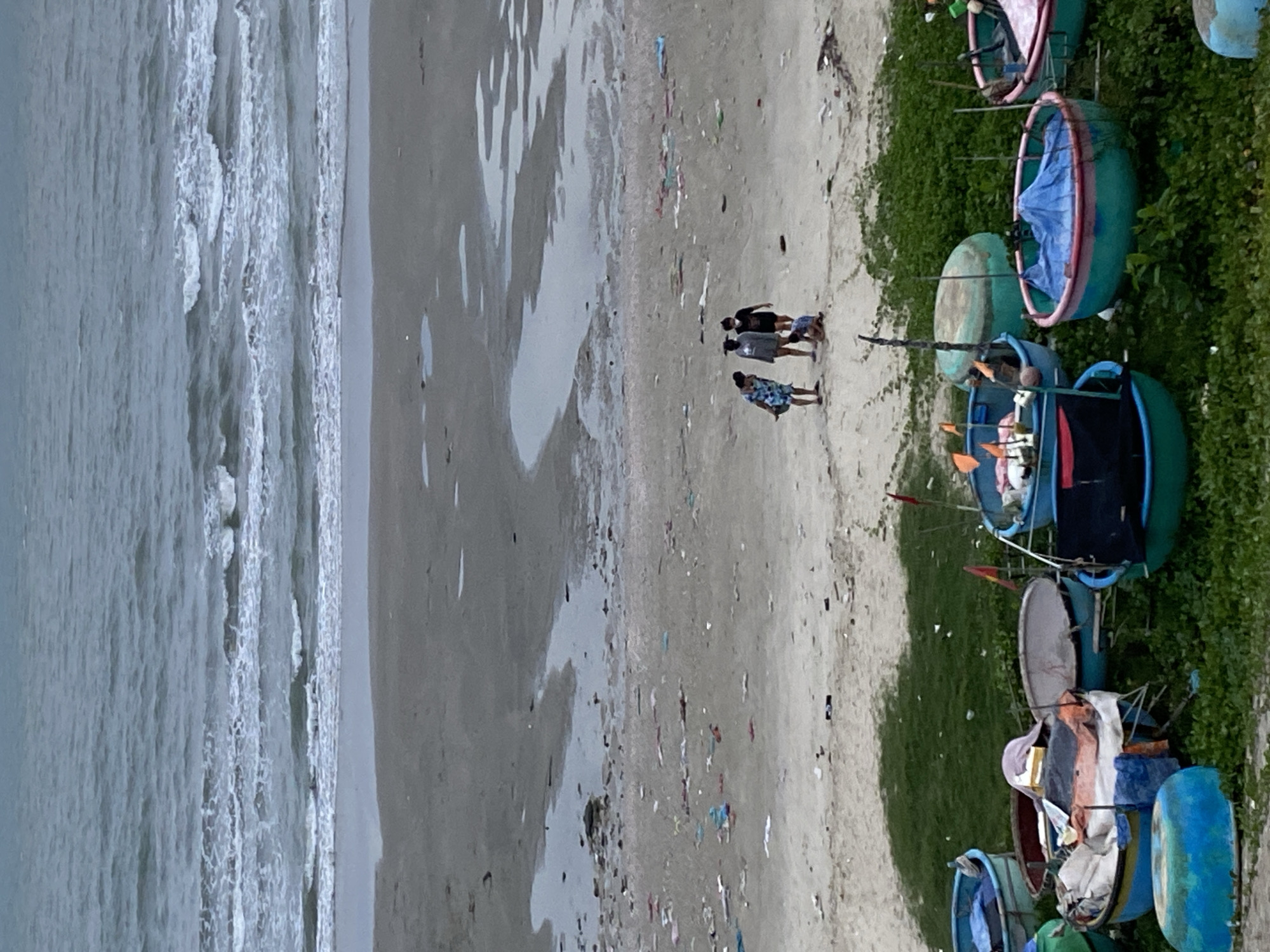 This media file depicts a beach scene in Mũi Né, Vietnam. The image shows a stretch of sandy beach with the ocean on the left and a line of round, basket-like boats on the right, nestled amongst green vegetation. A few people are visible walking on the wet sand near the water's edge. The boats are colorful, with green exteriors and pink or blue rims, and some are filled with various items like fabric and equipment. The water appears to be a muted grey-blue, with waves breaking along the shore. The sand is littered with small pieces of debris, suggesting it may not be entirely pristine. The lighting suggests it is daytime, and the weather appears overcast or hazy. The overall impression is a quiet, coastal setting with a focus on the traditional fishing boats characteristic of the region. No text is visible in the image.