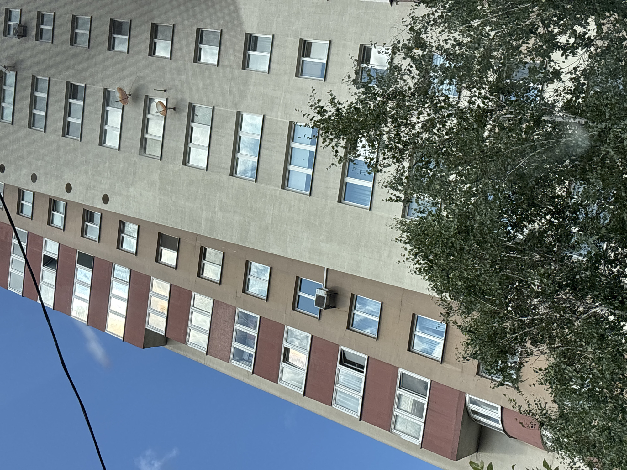 The image depicts a tall apartment building with a distinct facade, partially covered by lush green trees. The building is angled, suggesting it's viewed from below or at a slight angle. The upper portion of the building is a light tan color, while the lower sections feature a reddish-brown accent. Numerous windows are visible, some reflecting the blue sky and clouds, others showing the interiors with blinds or curtains. Two satellite dishes are mounted on the tan section of the building. An air conditioning unit is also attached to the exterior wall. The sky is a clear, bright blue with a few wispy white clouds. The trees in the foreground are dense and leafy, obscuring some of the lower floors of the building. The scene is characteristic of an urban environment in Minsk, Belarus, with its typical multi-story apartment blocks and interspersed greenery.