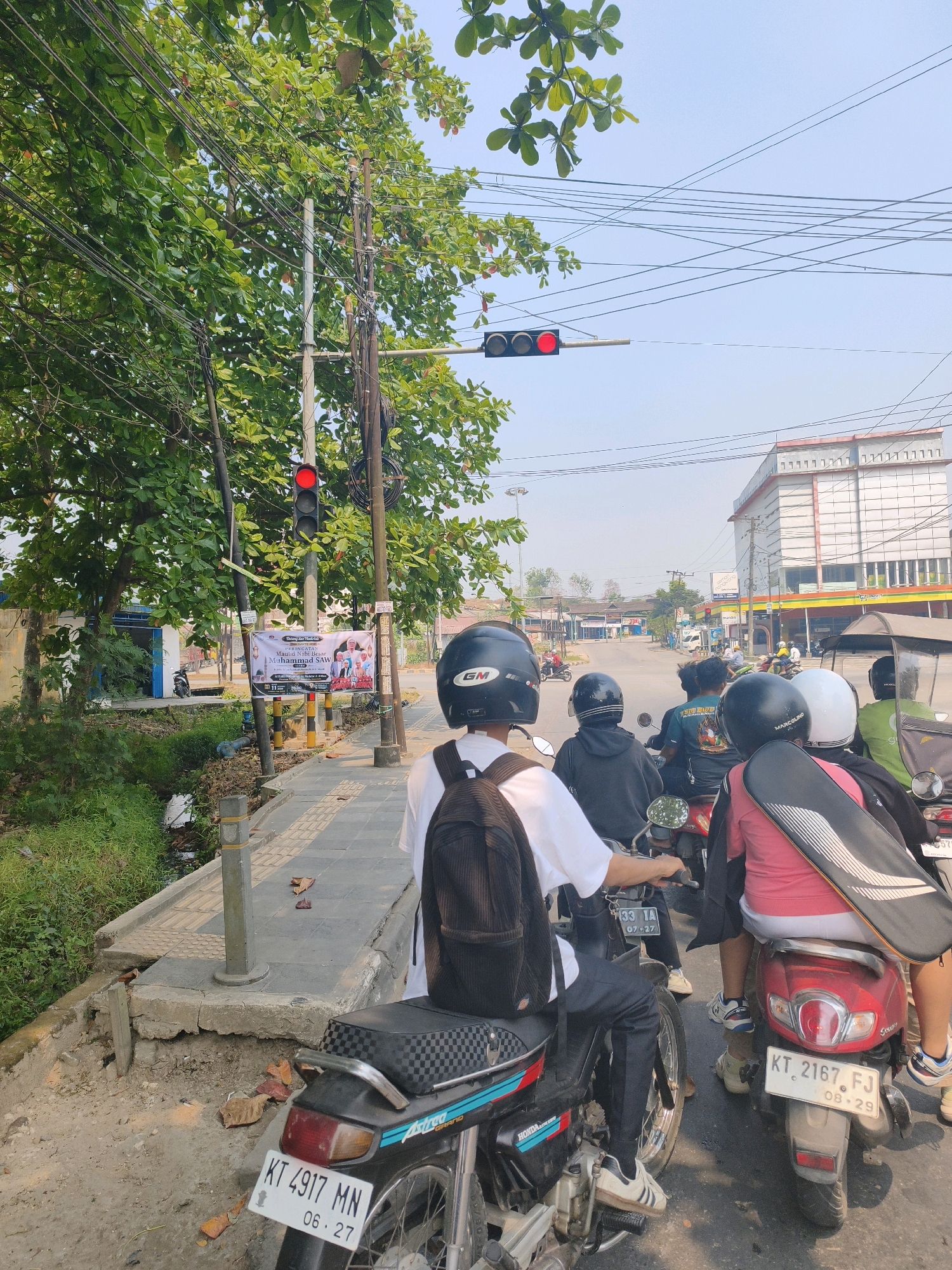 This is a street-level, eye-level, vertical, medium shot of a traffic jam on a sunny day in Samarinda, Indonesia. In the foreground, a motorcycle with the license plate KT 4917 MN, dated 06.27, is stopped at a red traffic light. The rider, wearing a black helmet and a white t-shirt, has a backpack on. Behind them, other motorcycles are also stopped, some with multiple passengers. The traffic light, which is red, hangs over the intersection, with a second traffic light also visible in the distance, also showing red.
The environment includes a sidewalk with tactile paving, a small, overgrown drainage ditch on the left, and lush green trees with visible branches and leaves overhead. Utility poles with numerous wires are present. In the background, there are several buildings, including a prominent multi-story white building with red accents and what appears to be a commercial establishment with yellow awnings. The road stretches into the distance, filled with more vehicles. A banner with text in Indonesian is attached to a pole on the sidewalk. The overall scene depicts a common urban traffic situation in Indonesia.
