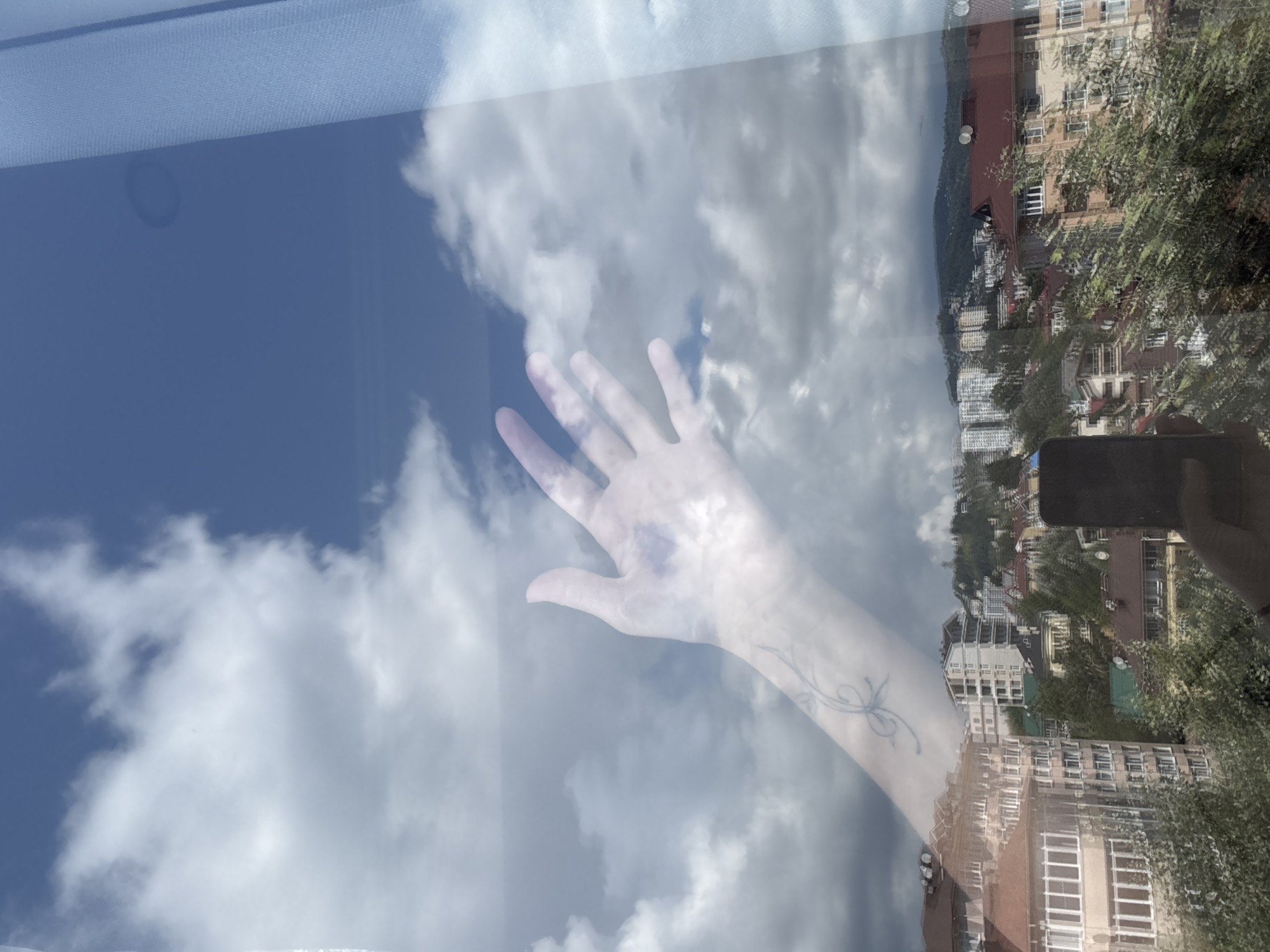 A reflection on a window shows a hand with a tattoo on the wrist reaching towards a cloudy sky. Below the sky, a city skyline is visible, with buildings of various heights and architectural styles nestled among green trees and hills. The scene appears to be set in a coastal city, possibly Sochi, Russia, given the lush vegetation and the style of the buildings. The weather is overcast, with dramatic clouds dominating the sky. The presence of the reflection suggests the photo was taken from inside a building looking out.