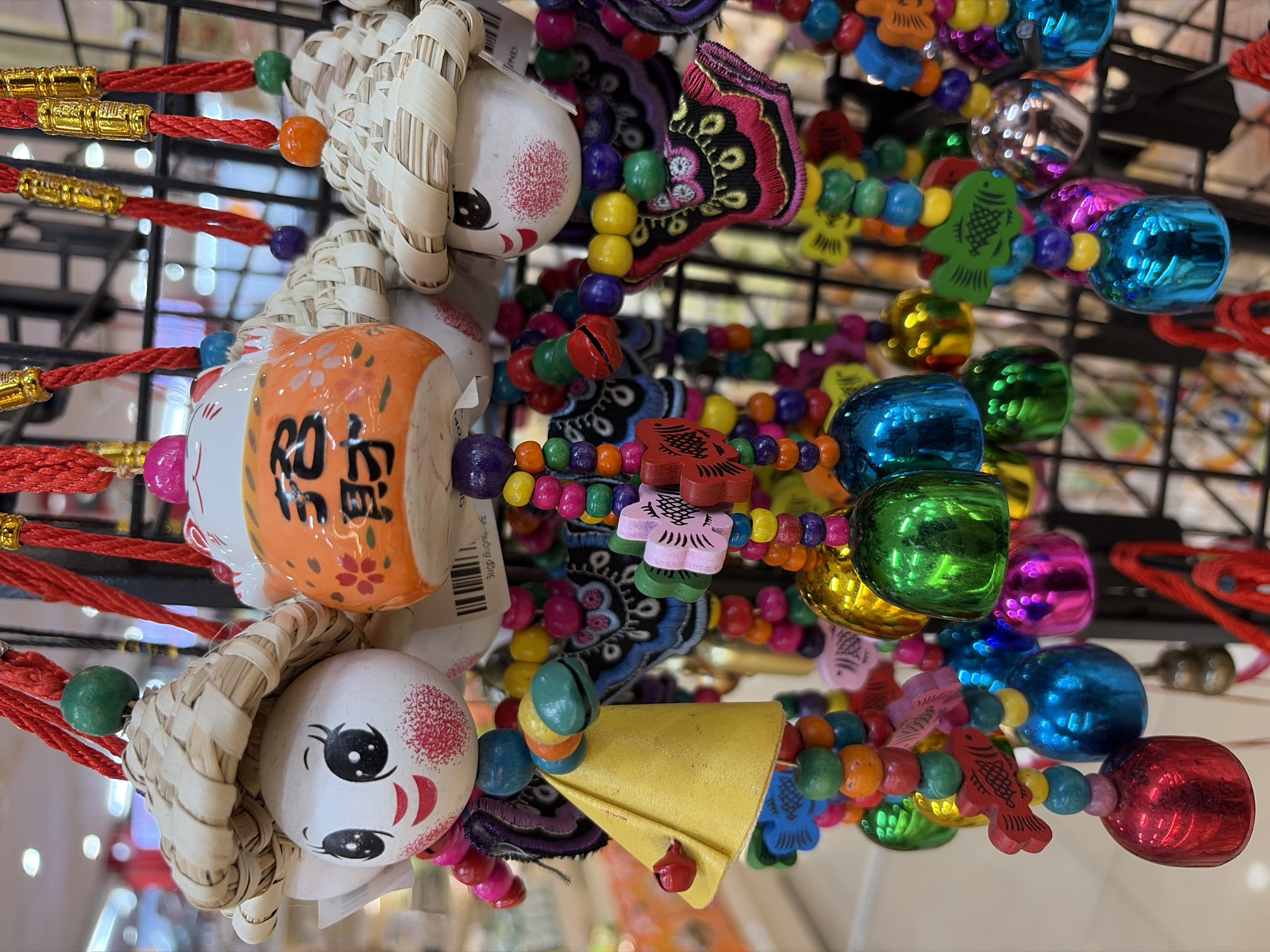 The image displays a close-up of numerous colorful hanging ornaments, likely souvenirs, displayed in an indoor setting. The ornaments are predominantly made of wood and metal, featuring a variety of shapes and designs. Several of the ornaments are shaped like dolls with straw hats and painted faces, while others are represented by intricately beaded strings adorned with small fish, flowers, and bell-shaped metal charms. The color palette is vibrant, with prominent use of red, green, blue, yellow, orange, and pink. The background appears to be a retail environment, with blurred shelves and other merchandise visible. The overall impression is one of festive and decorative items. There are no people visible in the image. The scene is likely from a market or souvenir shop in Đà Nẵng, Vietnam, given the style and commonality of such items in tourist areas there.