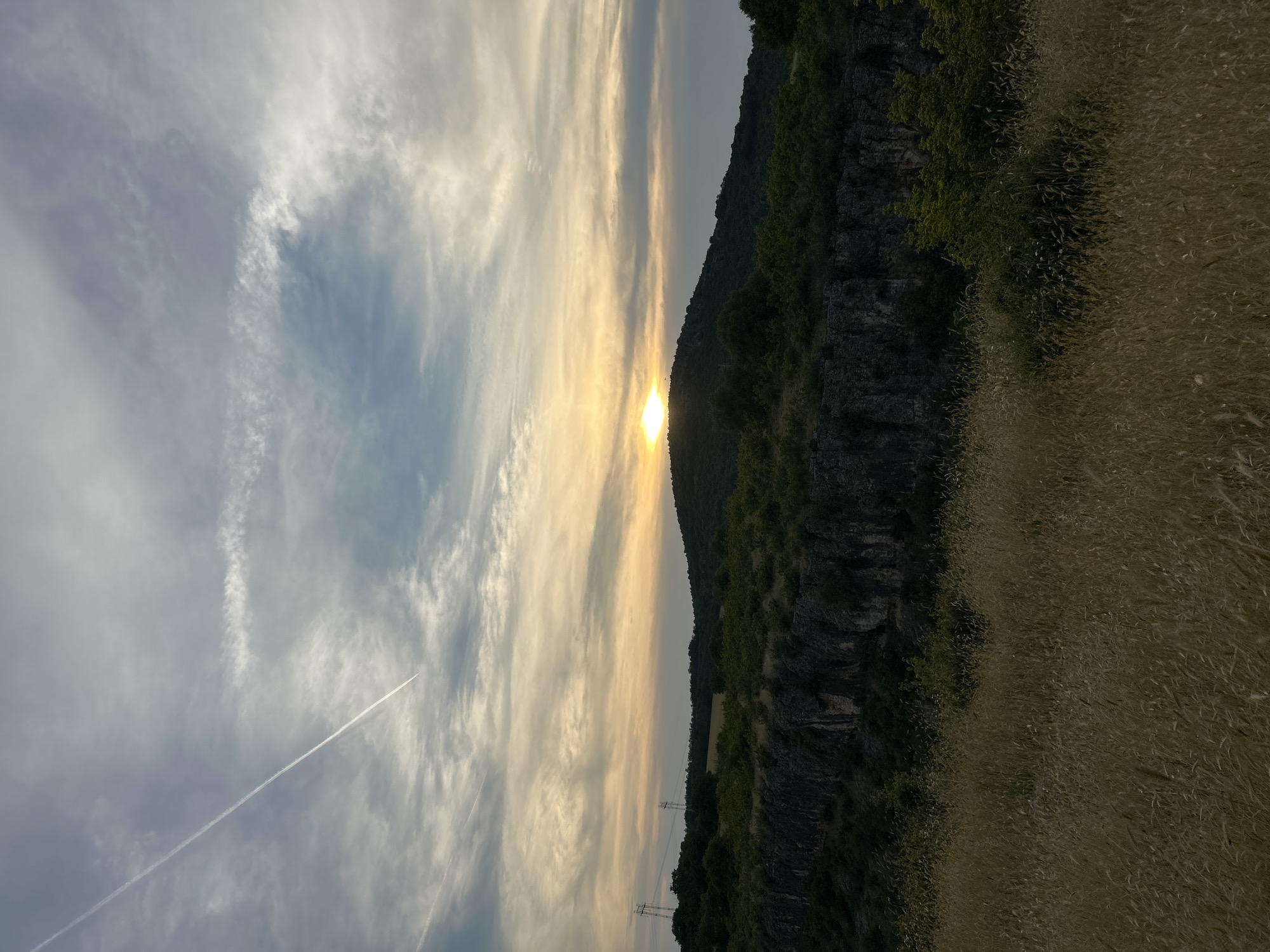 The image depicts a scenic sunset over a hilly landscape. In the foreground, tall, dry grass dominates the view, with its golden hues reflecting the fading sunlight. Behind the grassy field, a dark, rocky cliff face rises, covered in patches of green vegetation. Beyond the cliff, a rolling hill covered in dense green trees forms the middle ground, with a peak silhouetted against the sky. The sky itself is a canvas of dramatic clouds, streaked with wisps of white and tinged with the warm colors of sunset, including a bright yellow sun just beginning to dip below the horizon. A faint contrail from an airplane is visible diagonally across the upper left portion of the sky. In the distance, two tall power line pylons are visible on the left side of the frame. The overall impression is one of natural beauty and tranquility.
This scene is characteristic of the area surrounding the Hotnitsa Waterfall and the valley of the Beli Lom River in Hotnitsa, Bulgaria, known for its picturesque natural formations and vast expanses of rolling hills and forests.