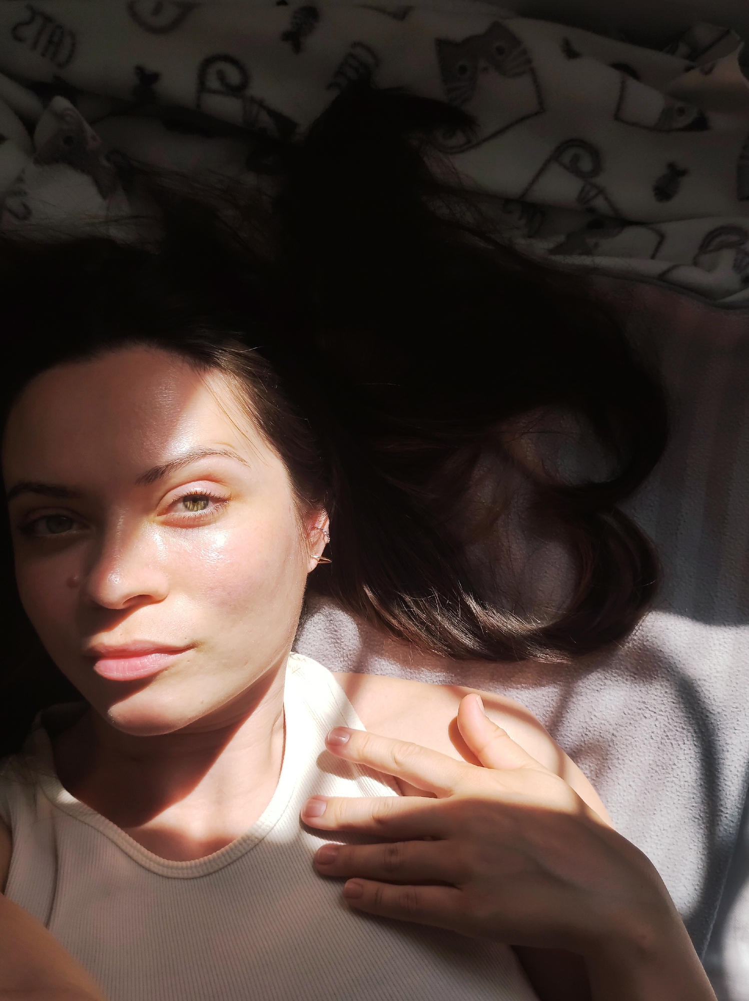 A young woman with long, dark hair is lying on her back on a bed. She is wearing a white tank top. The room is illuminated by sunlight, casting shadows across her face, chest, and hand. The bed is covered with a light-colored blanket with a repeating pattern of cats and fish. The location cannot be determined as Chaikovsky, Russia, as there is no indication of this within the image.