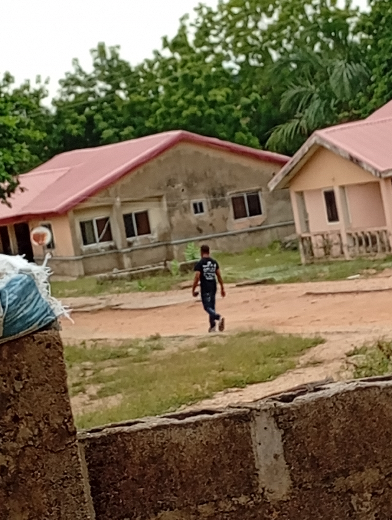 The image depicts a dirt ground landscape with two buildings and a person walking in the middle. The buildings are simple, one-story structures with red roofs, light-colored walls, and visible windows. Green trees are visible in the background, suggesting a natural environment. A person is walking away from the camera. In the foreground, there is a portion of a concrete structure, possibly a wall or fence. The location appears to be in a rural or semi-rural area. The location details provided are "Unknown, Unknown".