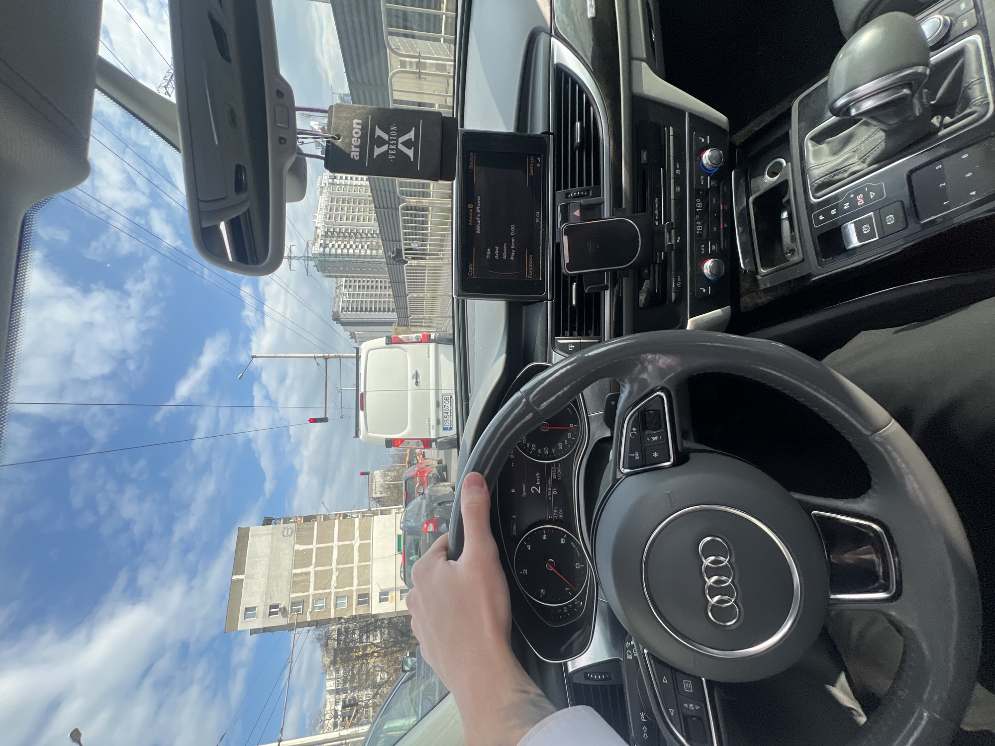 The scene is captured from the driver's perspective inside an Audi vehicle, which is stationary or moving at 2 km/h in traffic during daylight hours. The driver's left hand, wearing a white shirt cuff, rests on the upper left portion of the steering wheel, identifiable by the Audi rings logo. The instrument cluster shows an outside temperature of 17.5°C, a total mileage of 177,341 km, and the time as 16:04. The infotainment system displays "Mihail's iPhone" as the media source with a play time of 0:00. A smartphone is mounted vertically on the dashboard, and an "areon - Version X" air freshener hangs from the rearview mirror. Climate controls are set to 18.0°C.
Through the windshield, a line of traffic is visible, including a white Peugeot van with Bulgarian license plate CB 5407 OB (indicating Sofia). A red traffic light is visible ahead. To the immediate left, a multi-story residential building with a light-colored facade, featuring a grid-like pattern and a distinctive "SMART" logo (visible in the crop) on its upper side, stands prominently. This building is located at **ul. "Montevideo" 18, Ovcha Kupel II, 1618 Sofia, Bulgaria.** In the background, to the right, a cluster of modern high-rise residential or commercial buildings can be seen. Overhead power lines, streetlights, and metal pedestrian or construction barriers line the roadside. The sky is partly cloudy blue.