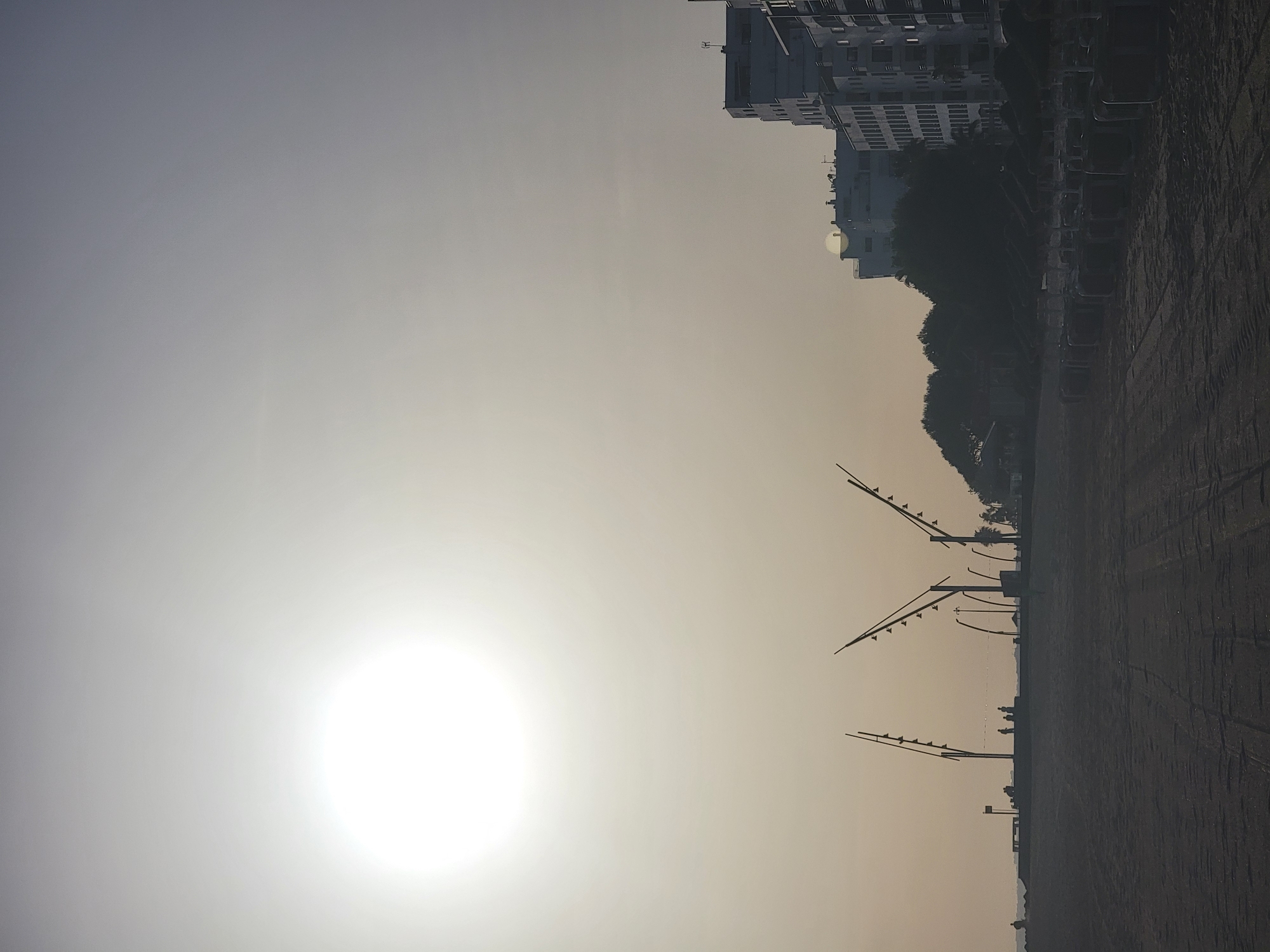 This is a vertical, wide-angle photograph taken at the beach in Quarteira, Portugal, during what appears to be either sunrise or sunset, indicated by the large, bright sun dominating the upper left quadrant of the sky. The sky is hazy and light, transitioning to a darker gray at the top.
In the foreground, a sandy beach stretches across the bottom of the frame, showing tire tracks and appearing damp in some areas. A row of empty beach chairs and umbrellas are neatly arranged on the sand in the middle right of the image, suggesting the beach is either just opening or closing for the day.
Further back, a concrete promenade or boardwalk runs horizontally across the mid-ground. Silhouettes of people are visible on this promenade, appearing as small, dark figures, some standing, others possibly walking or sitting. Several tall, modern lampposts with multiple lights are also silhouetted against the sky along the promenade, their distinctive designs adding a geometric element to the scene.
To the right of the promenade, a line of trees forms a dark, silhouetted band. Behind the trees and promenade, several multi-story buildings, characteristic of a coastal resort town, are visible. These buildings are lighter in color and appear to be hotels or apartment complexes, with visible windows and balconies. One building on the far right is particularly prominent, showcasing a modern architectural style. The overall atmosphere is calm and serene, with the bright sun and the muted colors of the hazy sky and sand creating a somewhat ethereal mood.