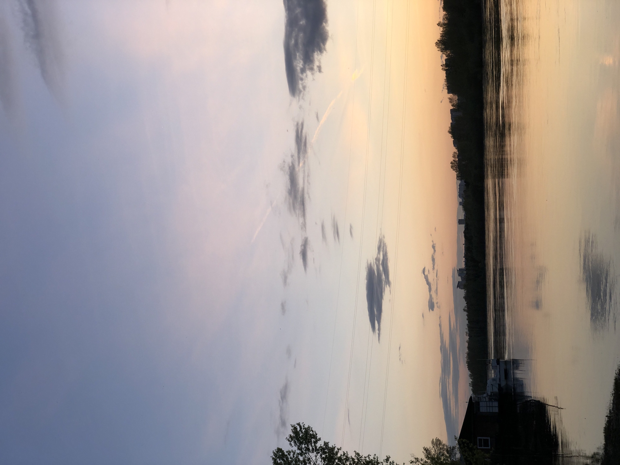 The image shows a sunset over a body of water, likely the Yenisey River in Krasnoyarsk, Russia, based on the visible cityscape and the river's prominence in the region. A building is present near the water's edge, and trees line the opposite bank. Power lines stretch across the sky, cutting through the gradient of colors transitioning from blue to orange and yellow. Clouds are scattered throughout the sky, and their reflections are visible in the water. The water surface is relatively calm, reflecting the colors of the sky and the silhouette of the trees.