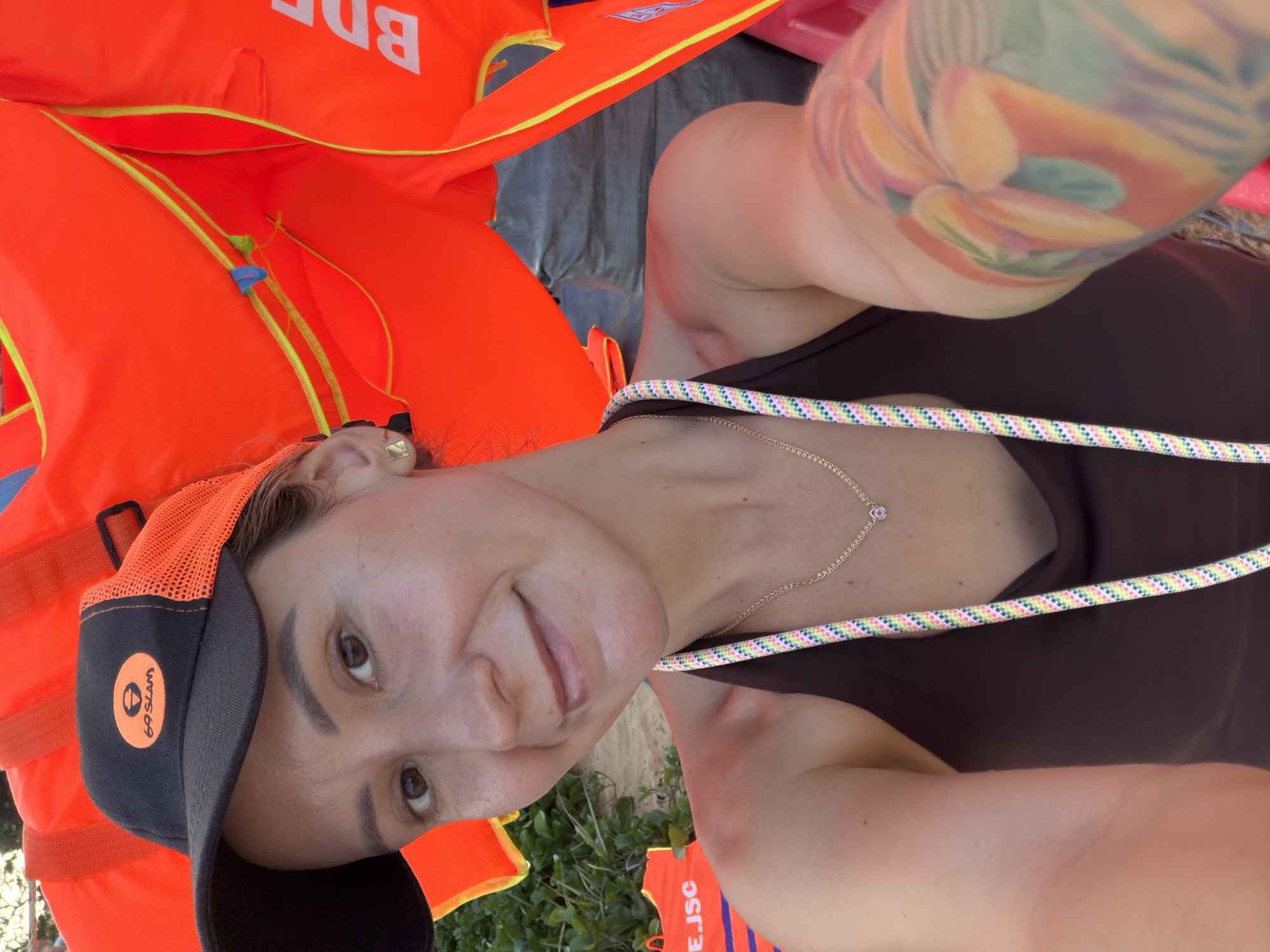 In a selfie taken outdoors, a woman with a dark baseball cap and a brown halter top smiles at the camera. The cap features an orange brim and a circular logo with the text "69 SLAM". She wears a delicate gold necklace with a small pendant and a colorful braided rope hangs across her chest. Her left arm, visible in the foreground, is adorned with a vibrant tattoo of tropical flowers. Behind her, bright orange life vests with yellow trim are stacked, and parts of what appear to be life vests with the text "BDE" and "EJSC" are also visible. The background suggests a beach setting, with sand and vegetation. The lighting indicates it is daytime.