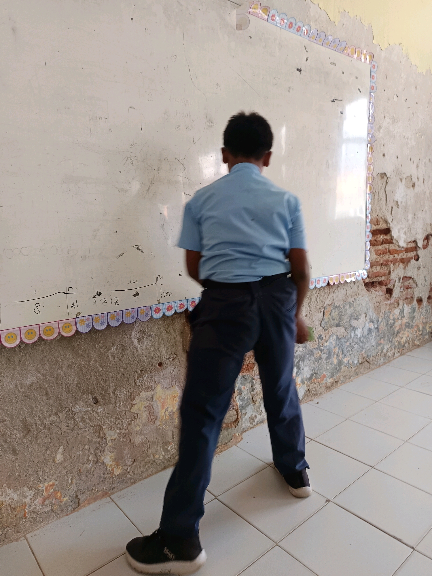A young male student wearing a light blue short-sleeved shirt and dark blue trousers stands facing away from the camera, in front of a grimy whiteboard. The whiteboard is decorated with a colorful border of cut-out paper images of flowers and smiley faces. The wall behind the whiteboard is crumbling, revealing exposed brickwork, and the floor is tiled with white squares. The overall environment appears to be a classroom in a state of disrepair, possibly located in Labuhan, Indonesia. The student is holding a green object in his right hand, the nature of which is unclear.