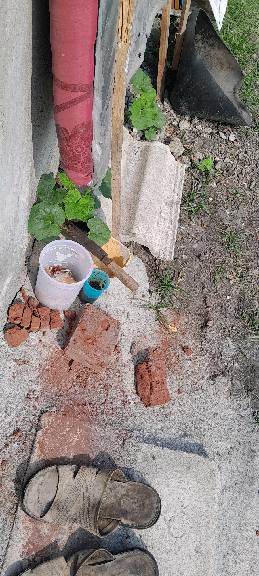 The image depicts a ground-level outdoor scene in Bendungan, Indonesia. In the foreground, a pair of worn, light-colored sandals are placed on a concrete surface. Scattered around the sandals and further up the image are pieces of broken red brick and brick dust. To the left, a white plastic cup contains some debris, possibly food scraps. Next to it, a smaller blue cup holds dark liquid. A large, old-fashioned knife with a wooden handle rests on a yellow shovel-like object. Behind these items, a bright red, rolled-up material, possibly a carpet or fabric, is leaning against a wall. Green leafy plants are growing around the base of this material. In the background, a black dustpan and wooden supports are visible, along with gravel and grass. The scene appears to be an outdoor storage or work area.