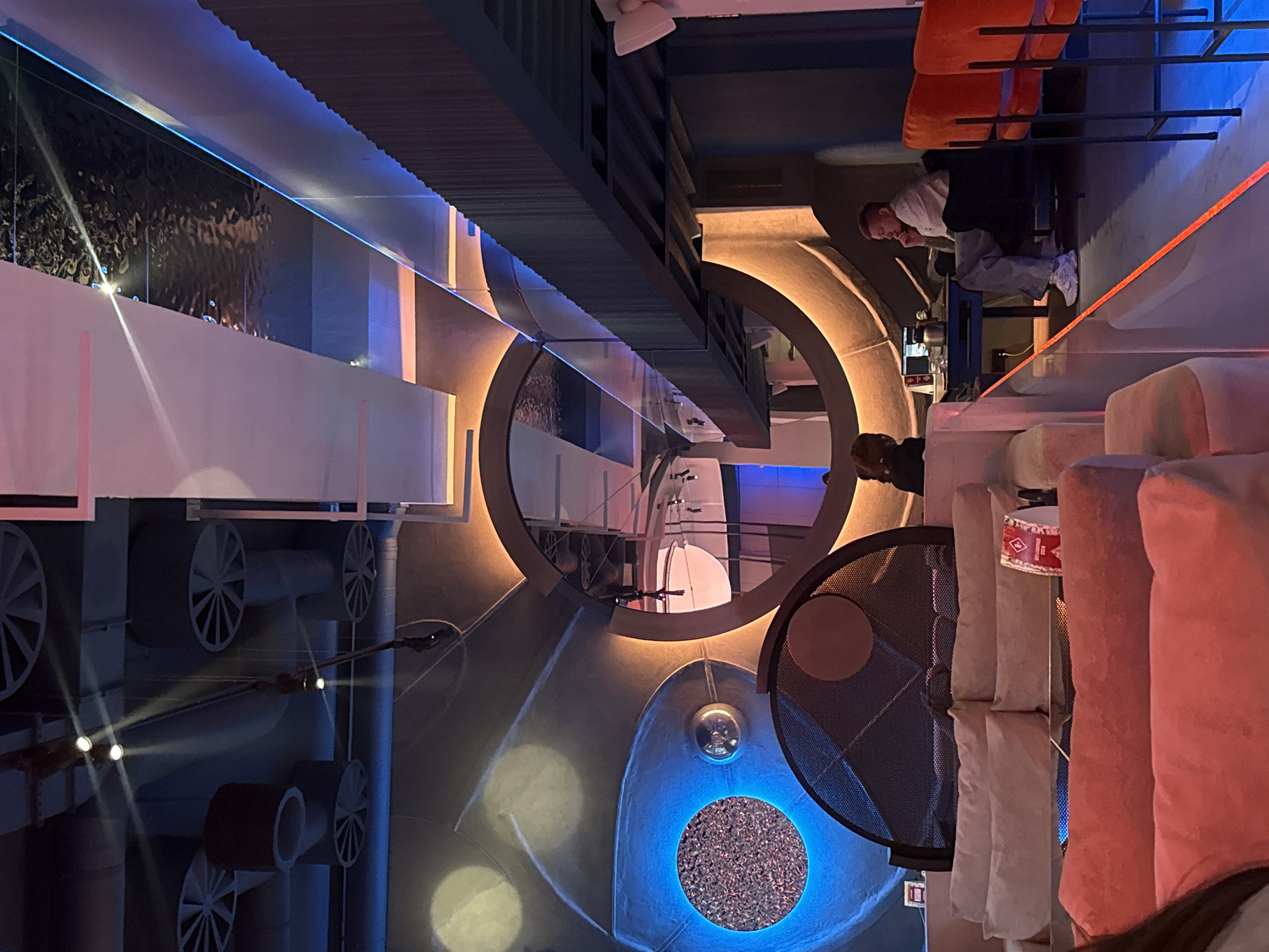 The image depicts the interior of a commercial space, likely a restaurant or bar, in Moscow, Russia. The space features a modern and industrial design. The walls have decorative elements, including circular light features and exposed ventilation systems. Seating is visible, with orange chairs and what appear to be decorative stacked cushions. A man is sitting at a high table, possibly waiting or working. A person is visible in the foreground. The lighting is a mix of warm and cool tones, creating an atmospheric ambiance. The overall setting appears to be a contemporary urban location.