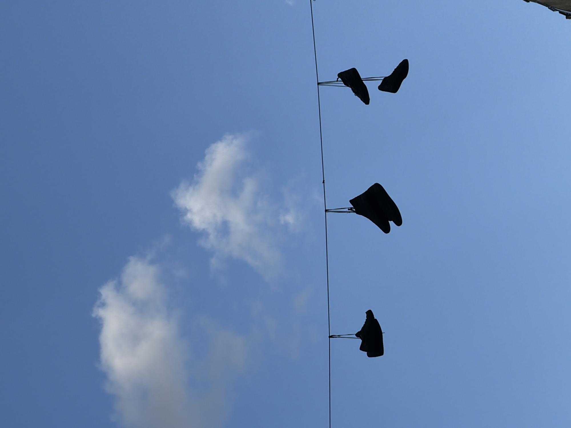 The image shows a low-angle view of three pairs of shoes hanging on a wire against a blue sky with some clouds. The shoes are silhouetted, making it difficult to identify their style or material. The wire is taut and runs vertically through the frame. It could be a clothesline, a telephone wire, or some other type of utility line. The location is unknown, but the practice of hanging shoes on wires is often associated with urban environments.