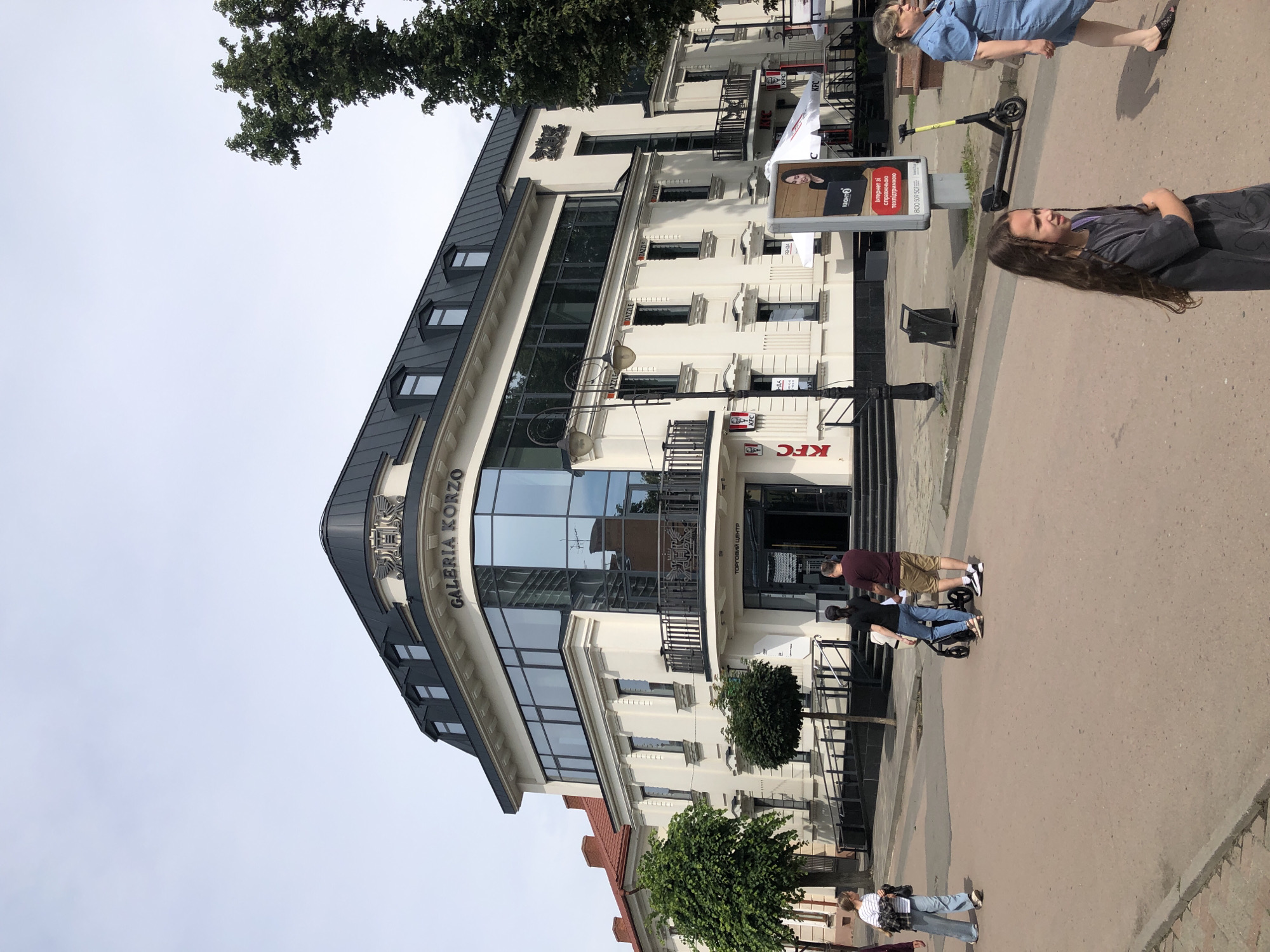 The image shows a street scene in Kolomyia, Ukraine, with the "Galeria Korzo" shopping center prominently featured. The building is cream-colored with a black mansard roof and large glass windows. The "Galeria Korzo" logo is visible on the upper facade, along with a decorative emblem. A KFC restaurant is located on the ground floor, with its signage clearly visible.
In the foreground, several people are walking on the paved street. A woman in a denim dress is walking from right to left. A young girl with long dark hair is standing and looking to the right, with her arms crossed. Further down the street, a couple with a stroller is walking away from the camera, and another person in light-colored clothing is walking from left to right. A yellow electric scooter is parked near a billboard advertising an internet service. The sky is overcast with a pale blue hue. Trees are visible on the left and right sides of the building.