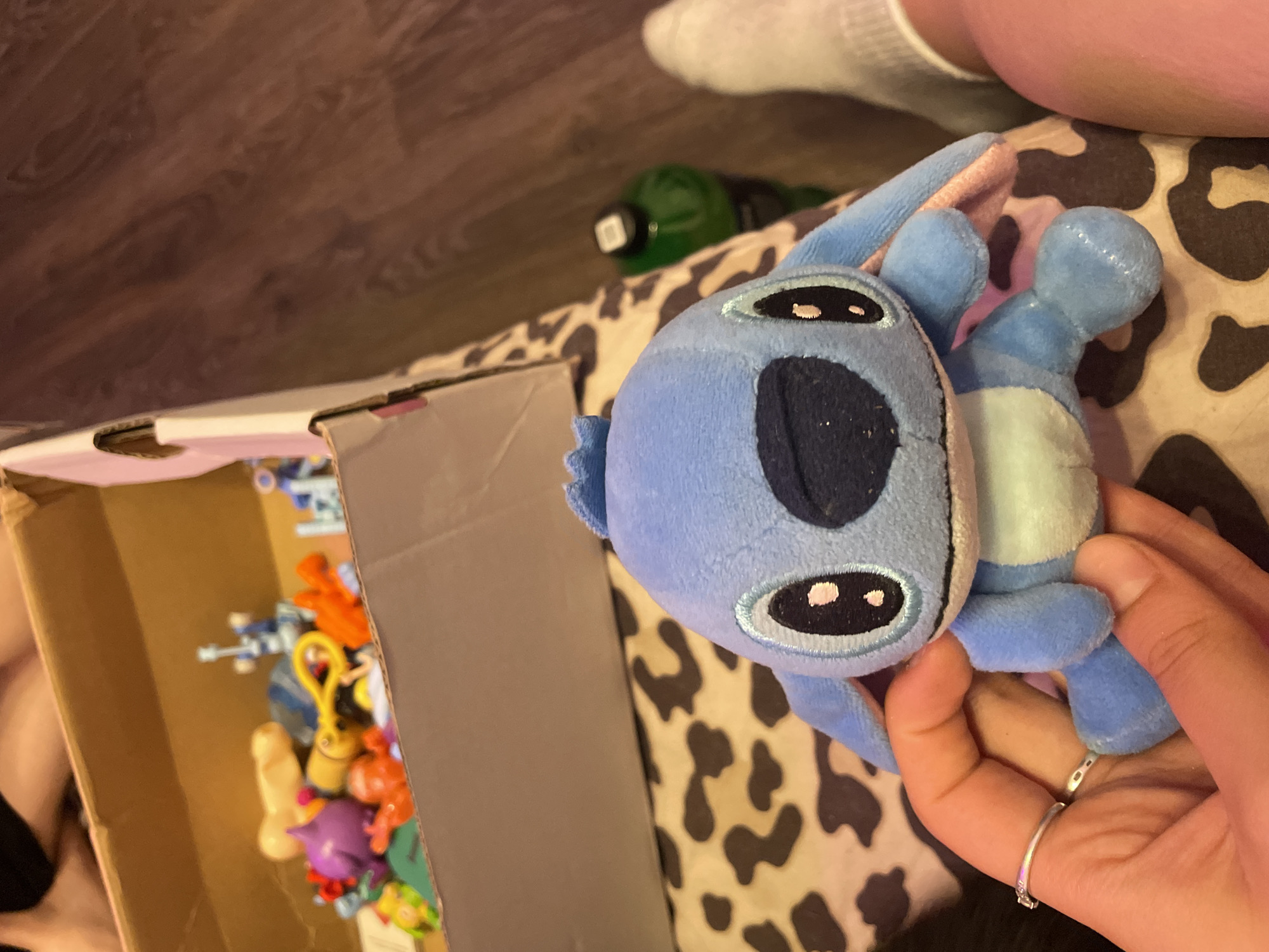 A light blue, plush toy resembling the character Stitch is being held in a person's hands. The toy has large black eyes with pink accents, a dark blue nose, and floppy ears. A silver ring with a small gem is visible on the person's left ring finger, and another plain silver ring is on their middle finger. In the background, a cardboard box filled with various colorful toys is visible. The box is placed on a surface with a leopard print pattern. To the right of the box, a green bottle and a person's lower leg wearing a white sock are visible. The floor appears to be made of dark wood. The scene is likely an indoor setting, possibly a home, in the city of Vaganovo, Russia.