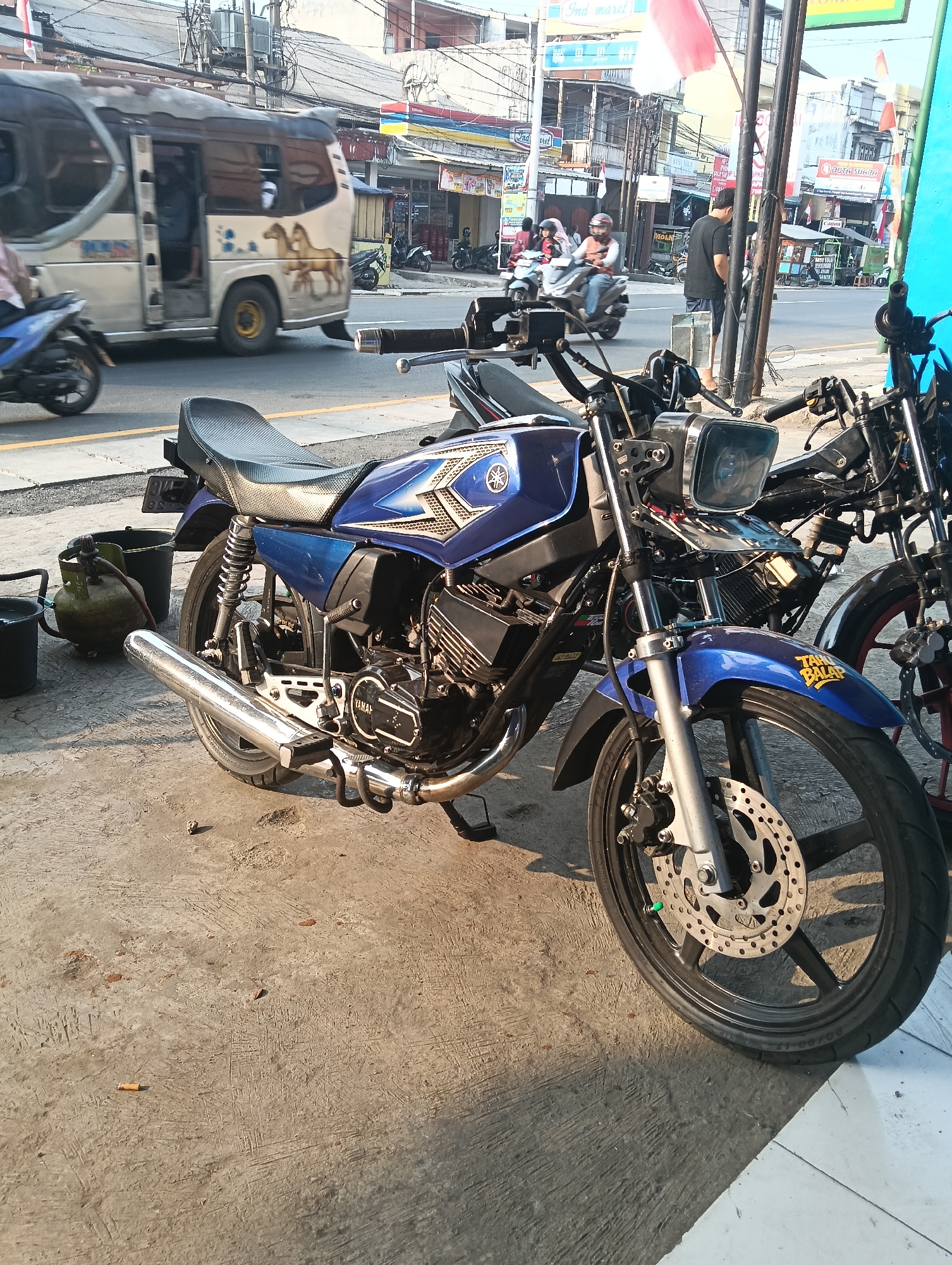 The image depicts a street scene in Desa Cipakat, Indonesia. A blue and silver Yamaha motorcycle, parked on a concrete sidewalk, is the primary subject. The motorcycle features a prominent exhaust pipe, a disc brake on the front wheel, and the Yamaha logo on its fuel tank. To the left of the motorcycle, a green gas cylinder and two buckets are visible on the sidewalk. In the background, a white van with horse decals is driving on the street, and other motorcycles and people are present. Buildings and shops line the street, suggesting a commercial or residential area. The overall environment is an outdoor urban setting with a sunny sky.